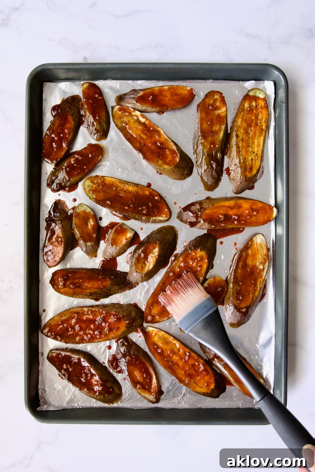 Slices of eggplant cut on the diagonal on aluminum foil-lined rimmed sheet pan being brushed with sauce.