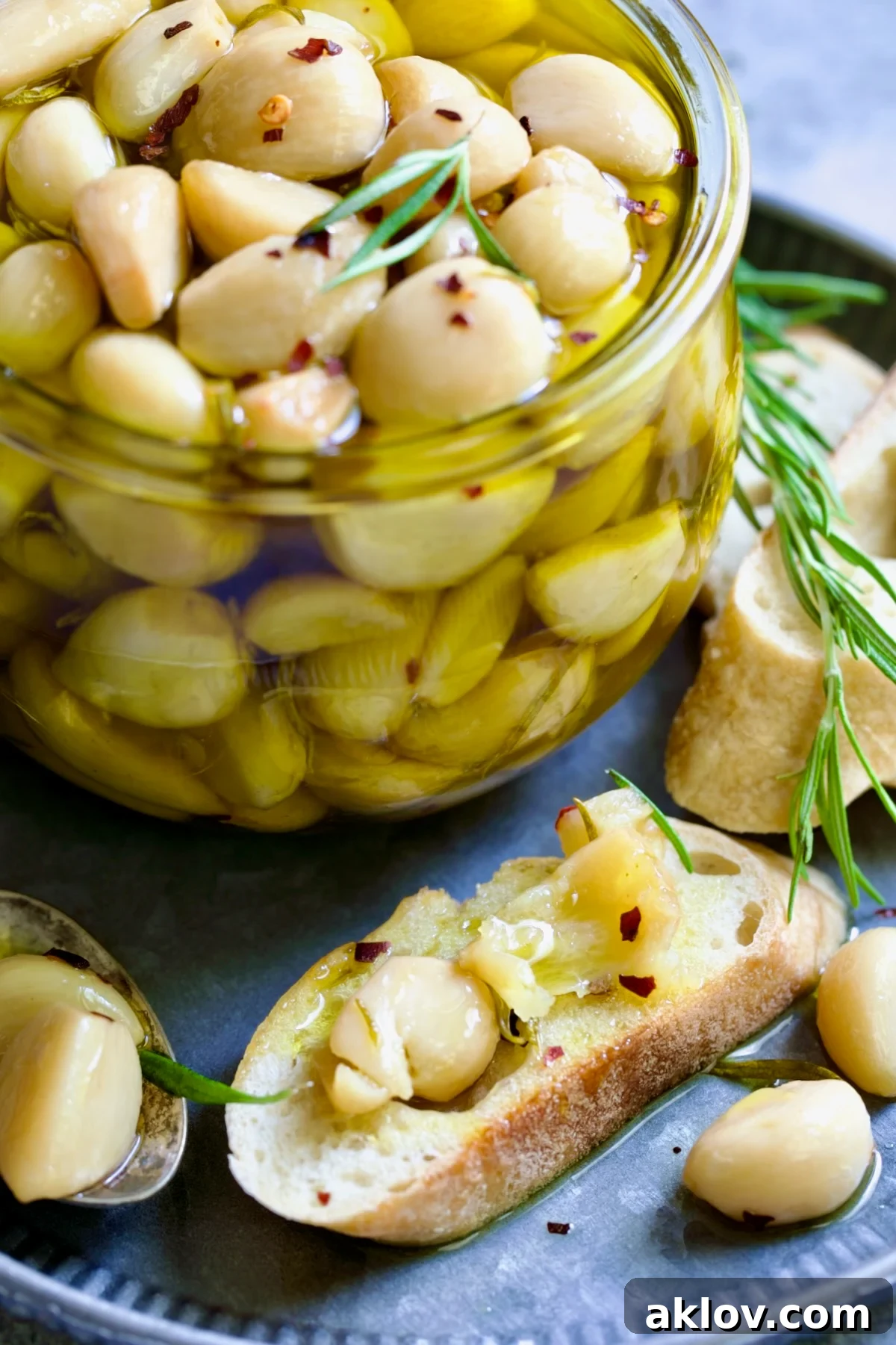A glass jar filled with golden garlic confit cloves and herbs, next to a piece of bread with mashed confit garlic.