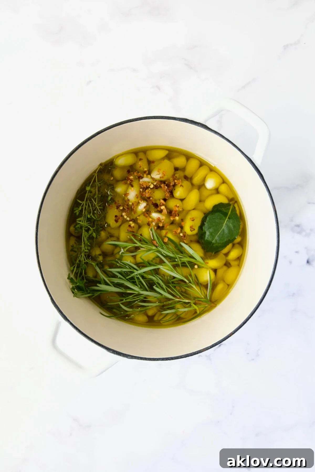 Peeled garlic cloves and fresh herbs submerged in olive oil in a white Dutch oven before being gently simmered.