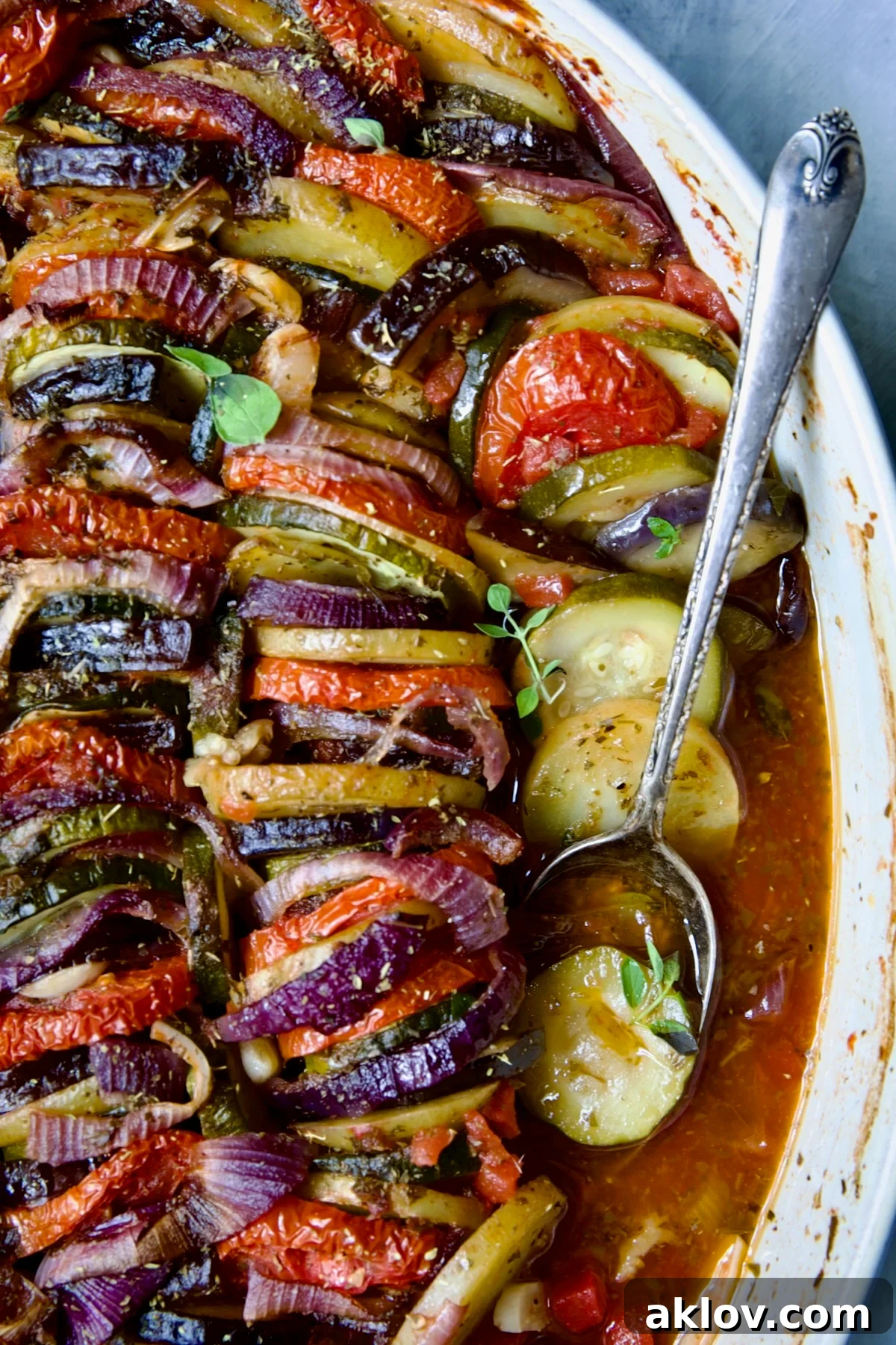 Briam (Greek Roast Vegetables) in oval white gratin dish with serving spoon exposing the tomato and olive oil juices.