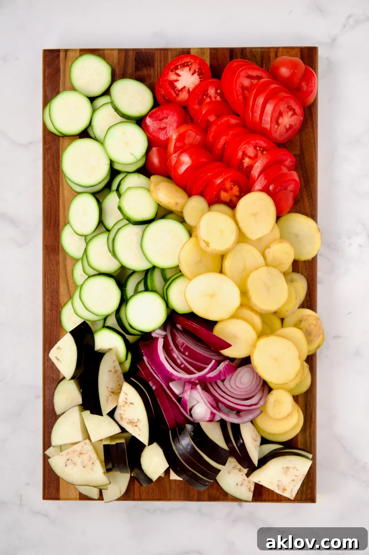 Sliced zucchini, potatoes, tomatoes, eggplant and red onion on wood cutting board.