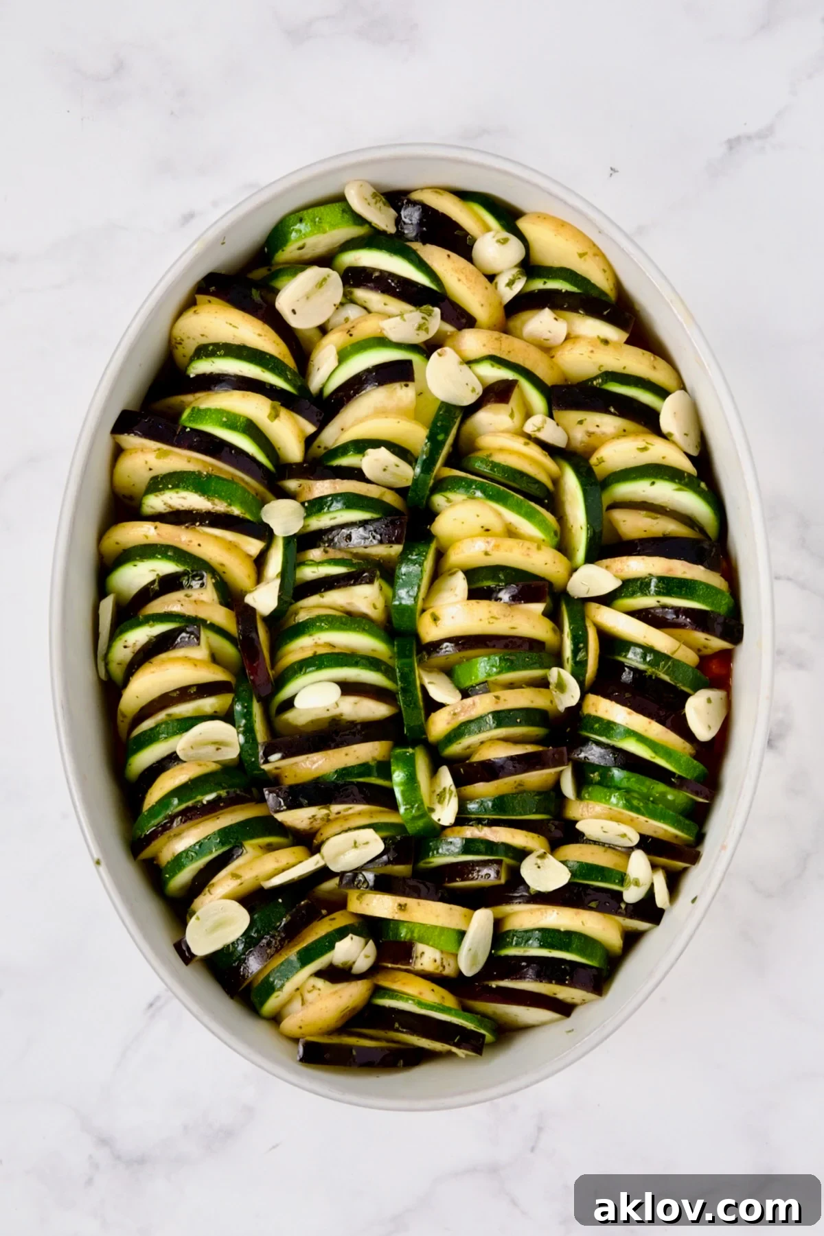 Potatoes, zucchini and eggplant arranged in oval white baking dish with sliced garlic over the top.