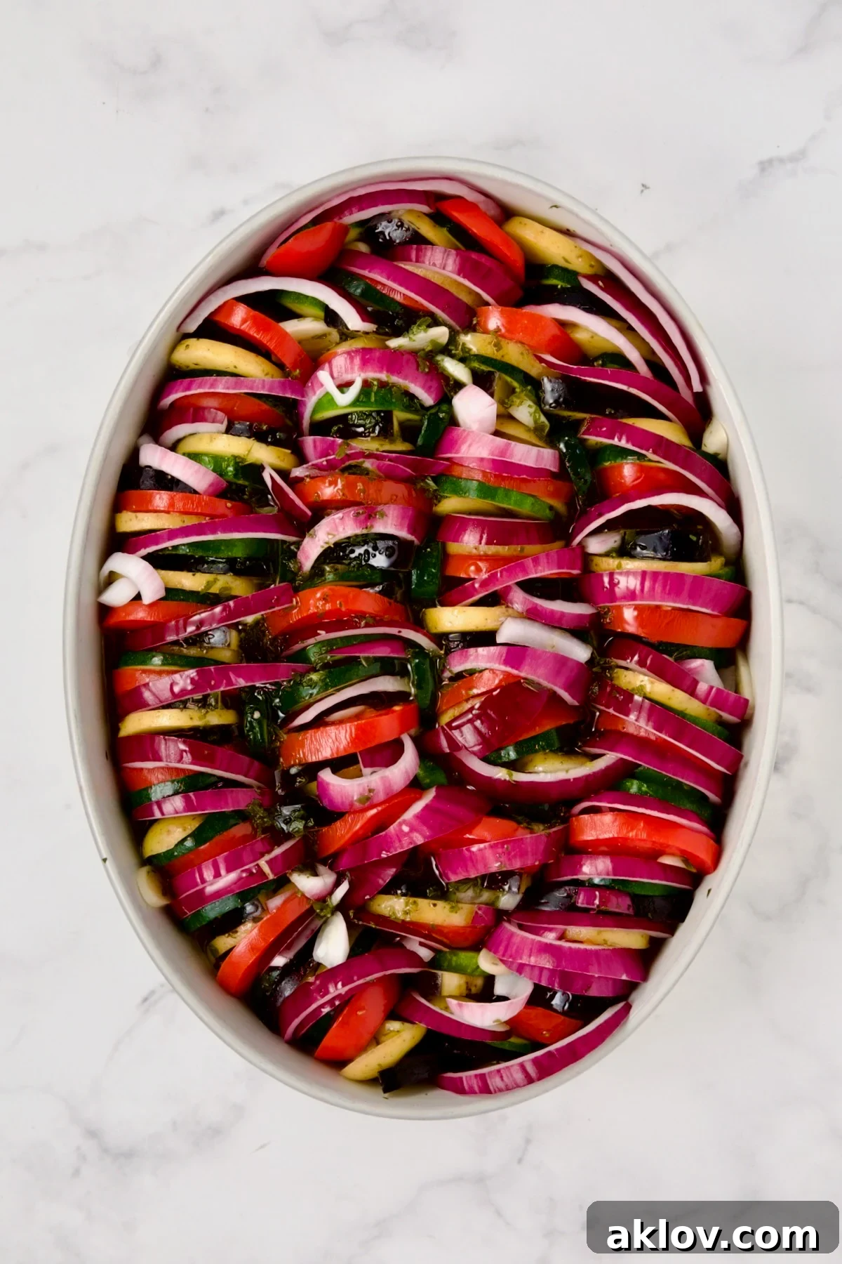 Red onion and tomatoes added to potatoes, zucchini and eggplant in oval white baking dish ready to bake.
