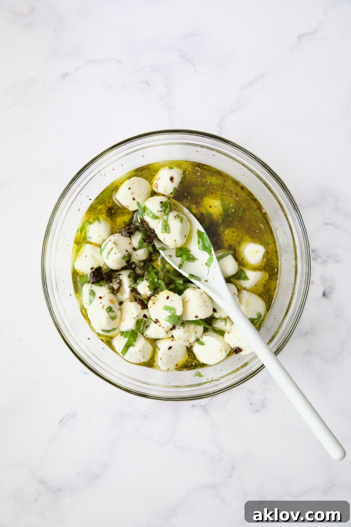 Fresh mozzarella balls and marinade being stirred with white spoon in glass bowl.