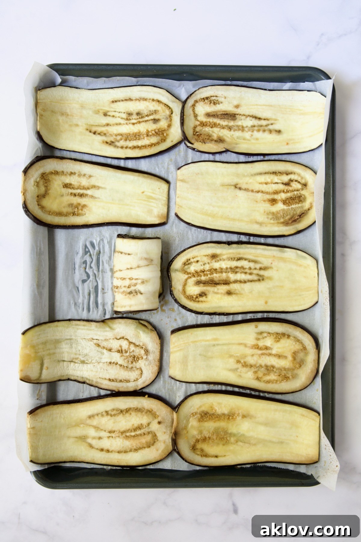 Ten baked eggplant slices on parchment paper-lined baking sheet with one rolled up to show pliability.