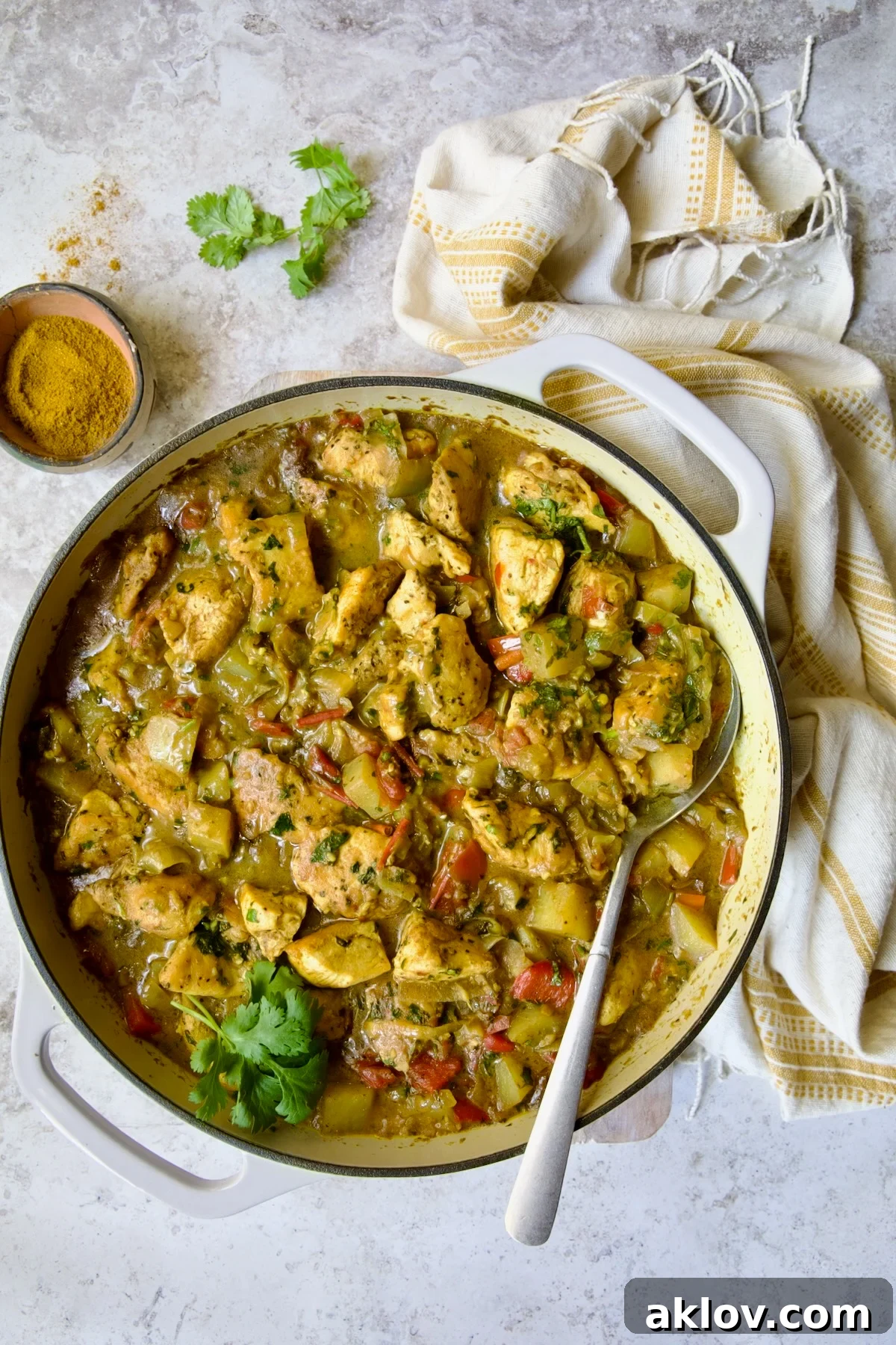 Comforting Chicken Potato Curry 14 A delectable Chicken and Potato Curry, rich and inviting, presented in a white enamel cast iron skillet, accompanied by a serving spoon.