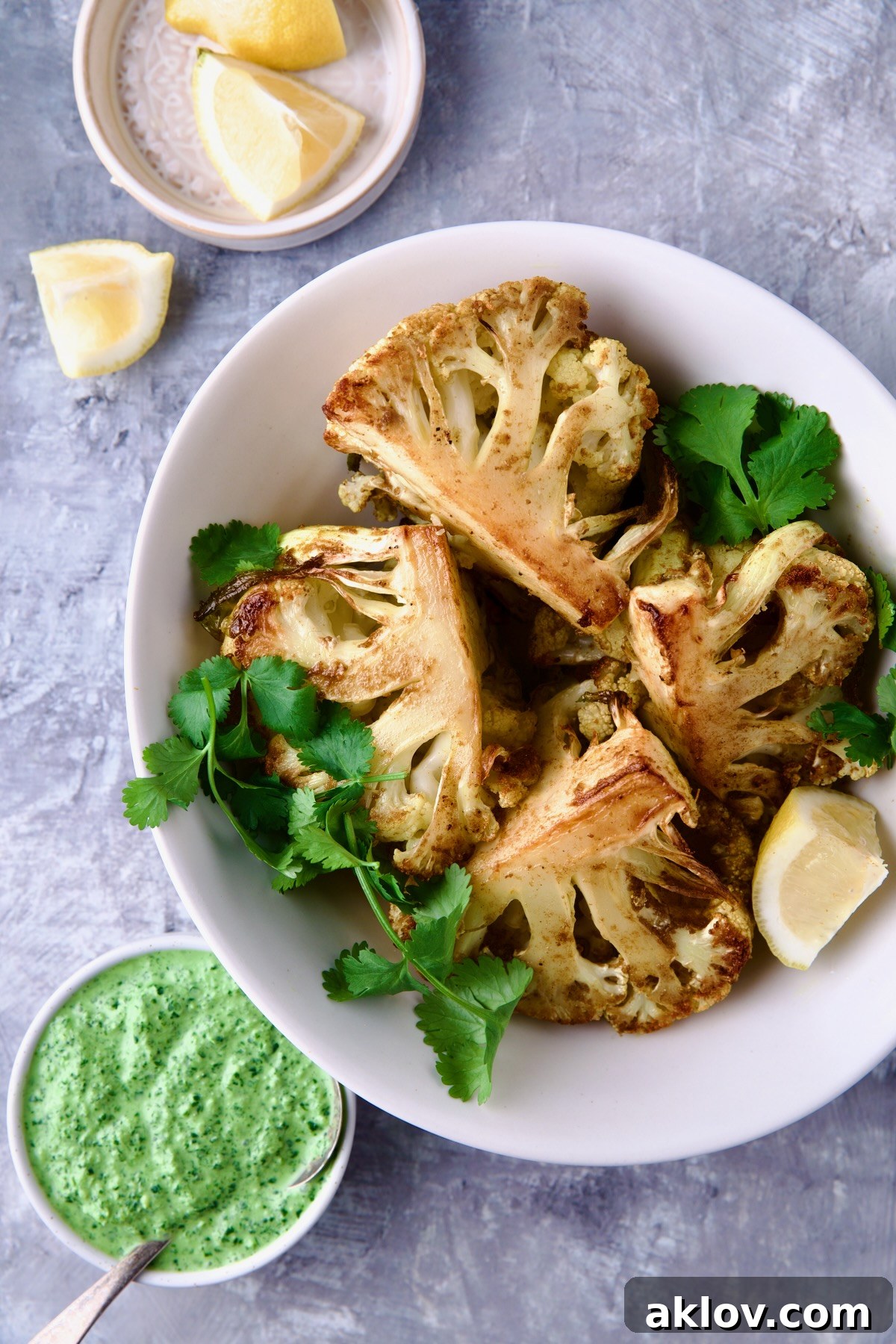 Four wedges of Curry Roasted Cauliflower in white oval bowl with Cilantro Chutney and lemon wedges.