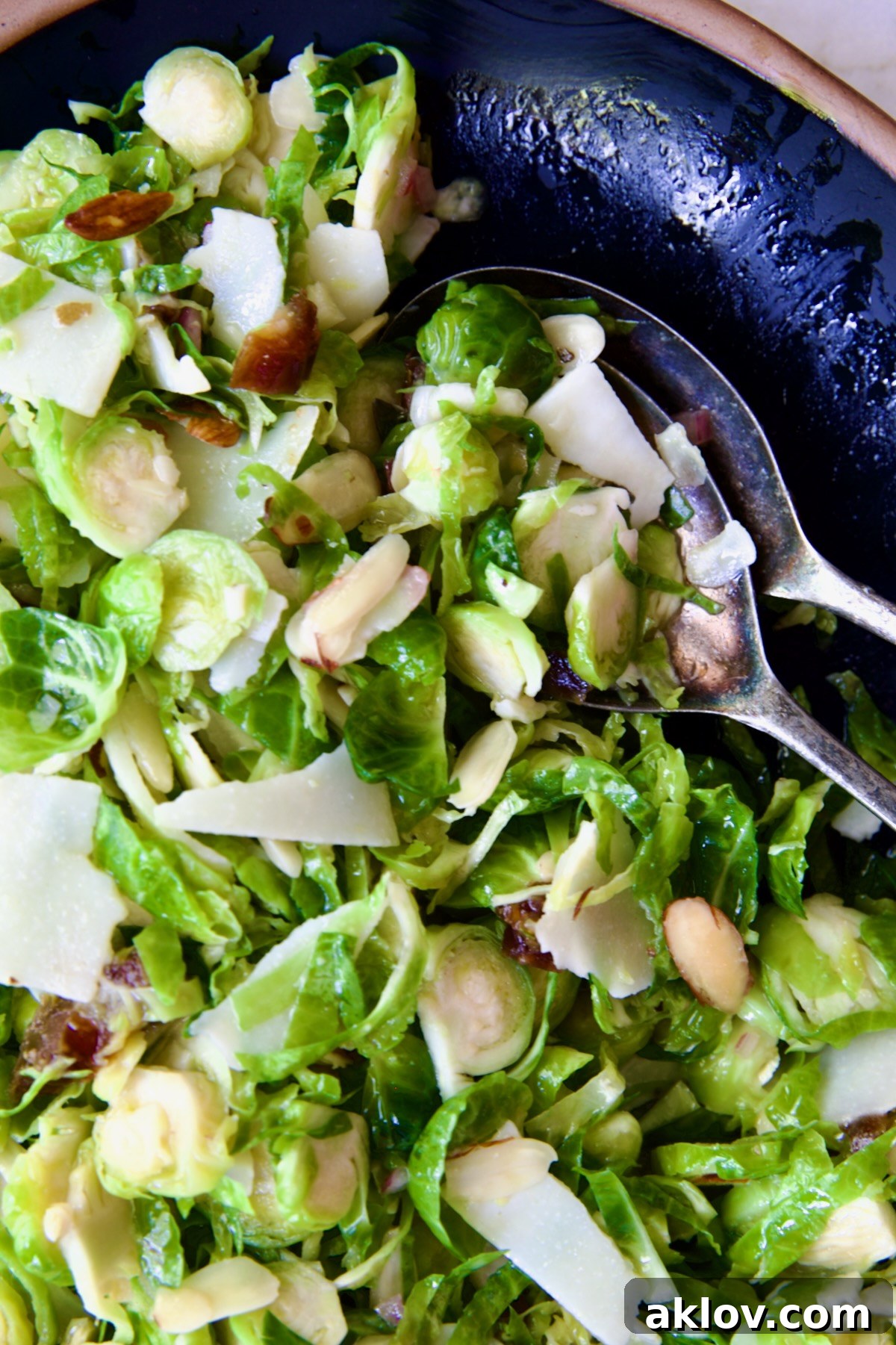 Zingy Shaved Brussels Salad 13 A generous serving of Shaved Brussels Sprouts Salad on a black platter with a brown rim, being scooped by serving utensils, inviting diners to enjoy its fresh flavors.