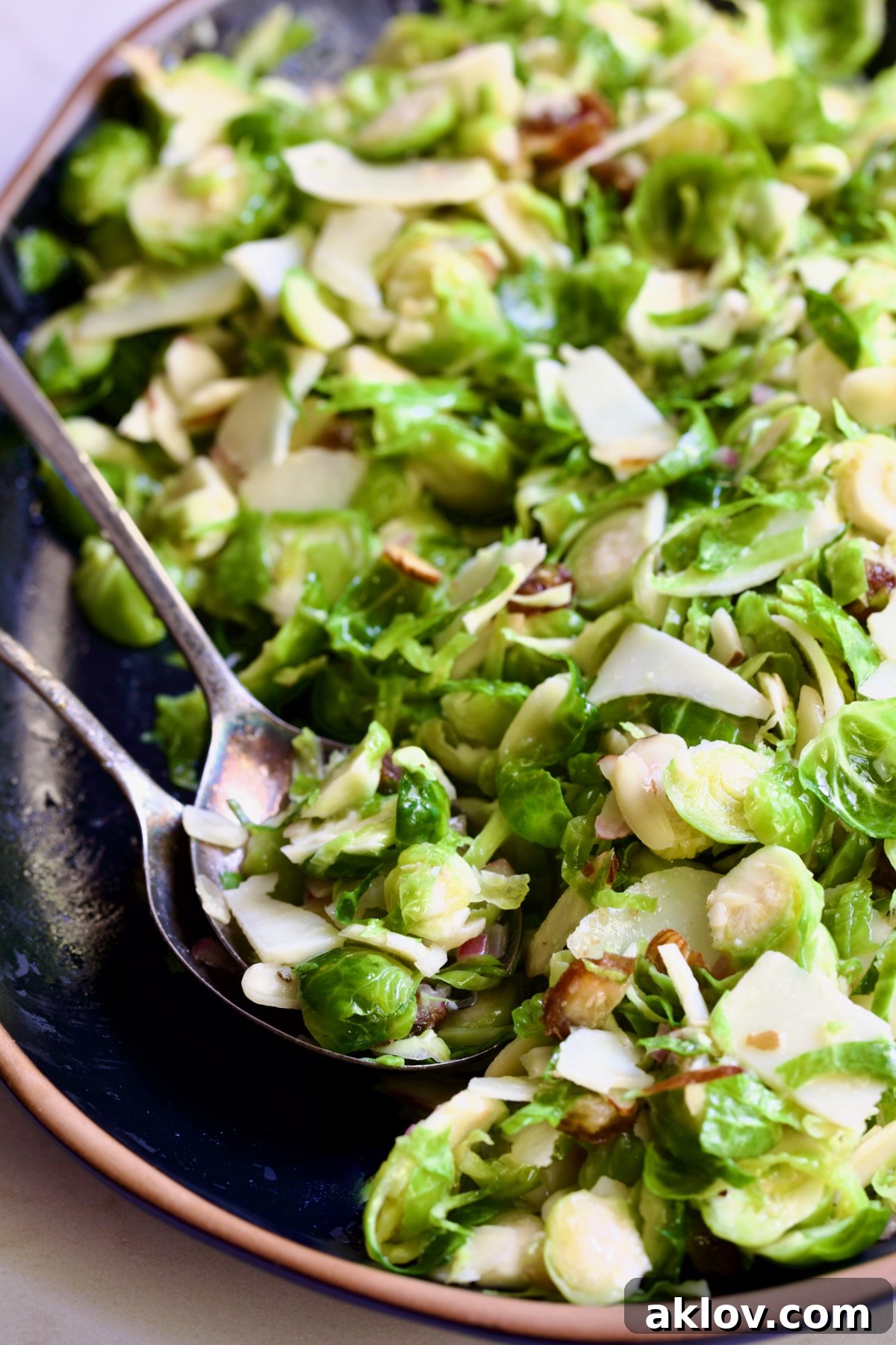 Zingy Shaved Brussels Salad 3 Serving utensils scooping a portion of Shaved Brussels Sprouts Salad from a black platter with a brown rim, highlighting its fresh ingredients and inviting texture.