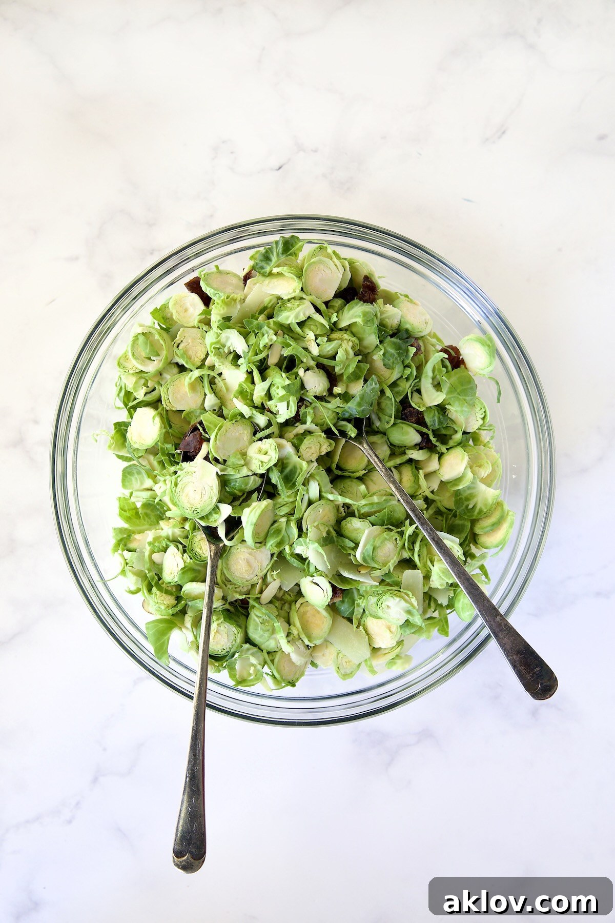 Zingy Shaved Brussels Salad 9 A vibrant mix of shaved Brussels sprouts, chopped dates, sliced almonds, and shaved Romano cheese being gently tossed together with serving utensils in a glass bowl, prior to the dressing.