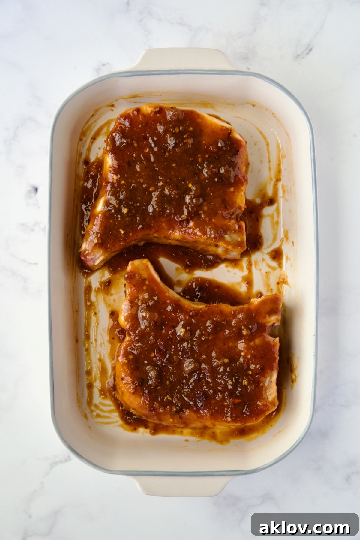 Two pork chops in a baking dish, fully coated with the apricot-chili crisp glaze on both sides, ready for the oven.