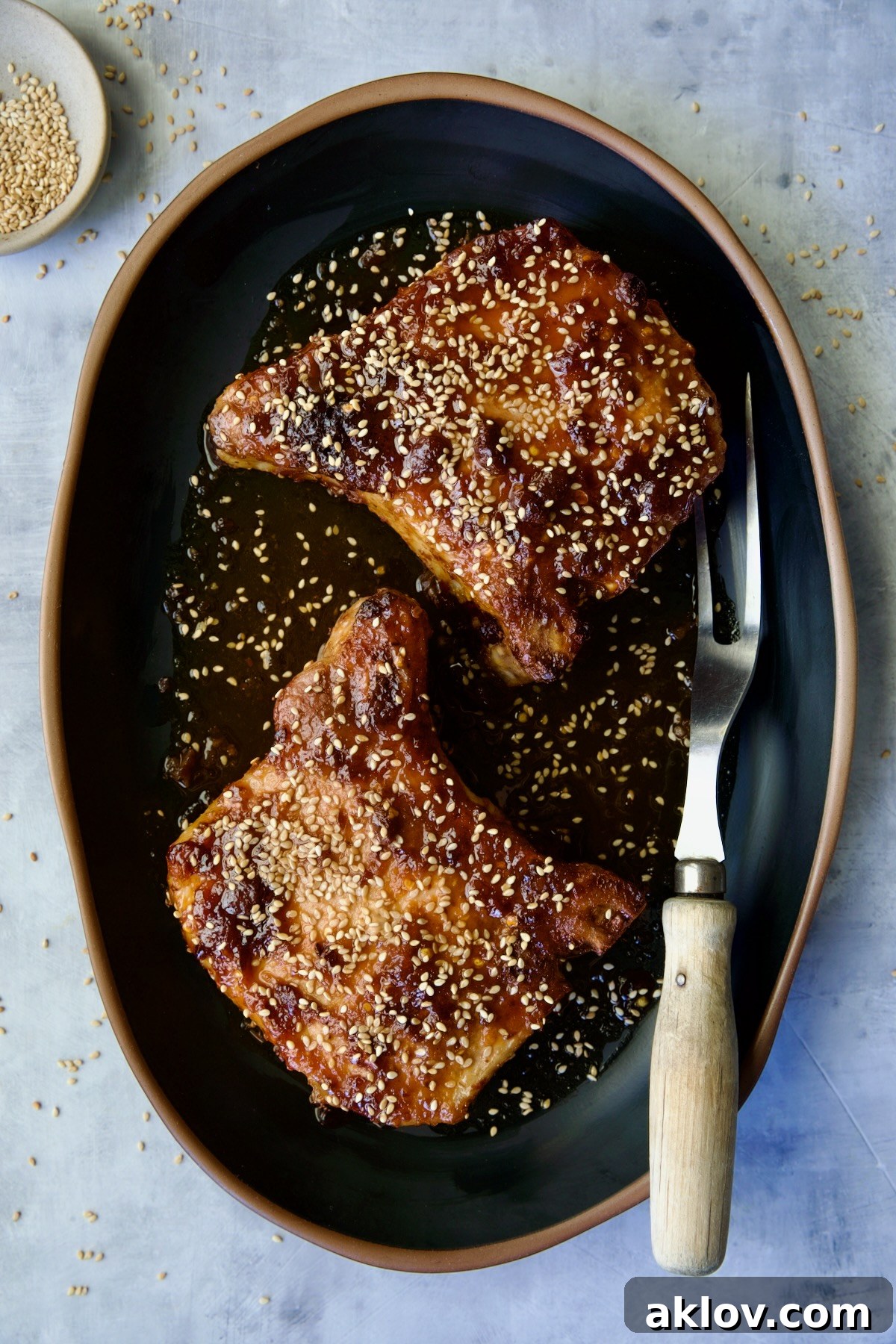 Two beautifully plated Asian Pork Chops on a sleek black oval platter, garnished generously with sesame seeds.