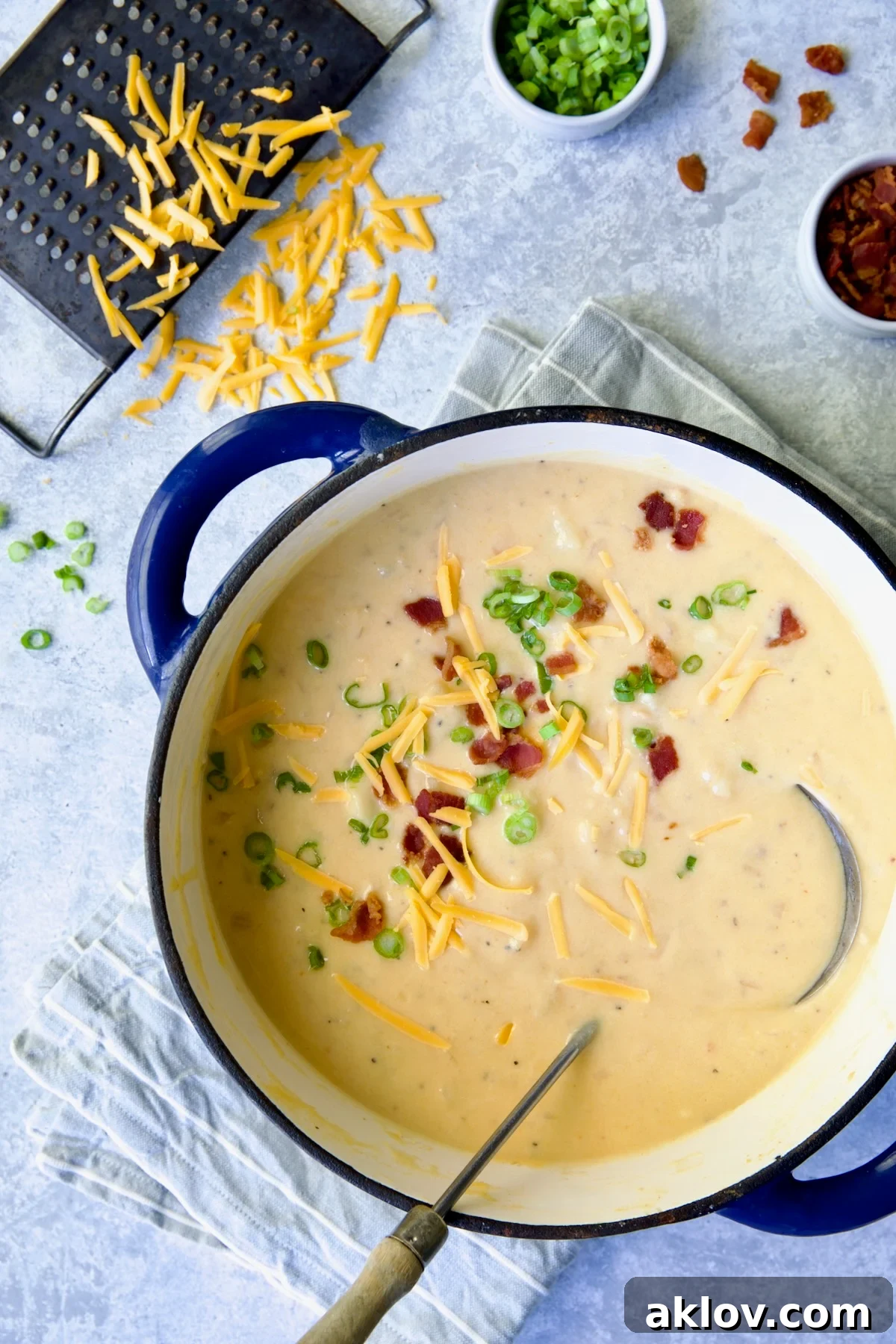 Cheddar Bacon Potato Soup in blue Dutch oven with soup ladle and cheese, scallion and bacon scattered about.