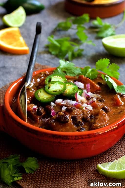 A rich, dark Feijoada (Brazilian Beef Stew) served in a rustic red clay bowl, elegantly garnished with vibrant jalapeno slices, fresh cilantro, and refreshing fruit wedges.