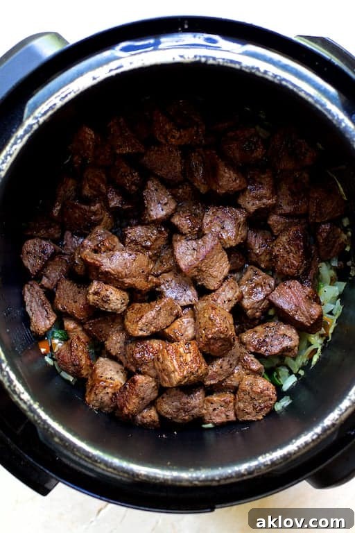 Browned beef cubes, alongside other ingredients, resting inside an electric pressure cooker before the lid is sealed for cooking.
