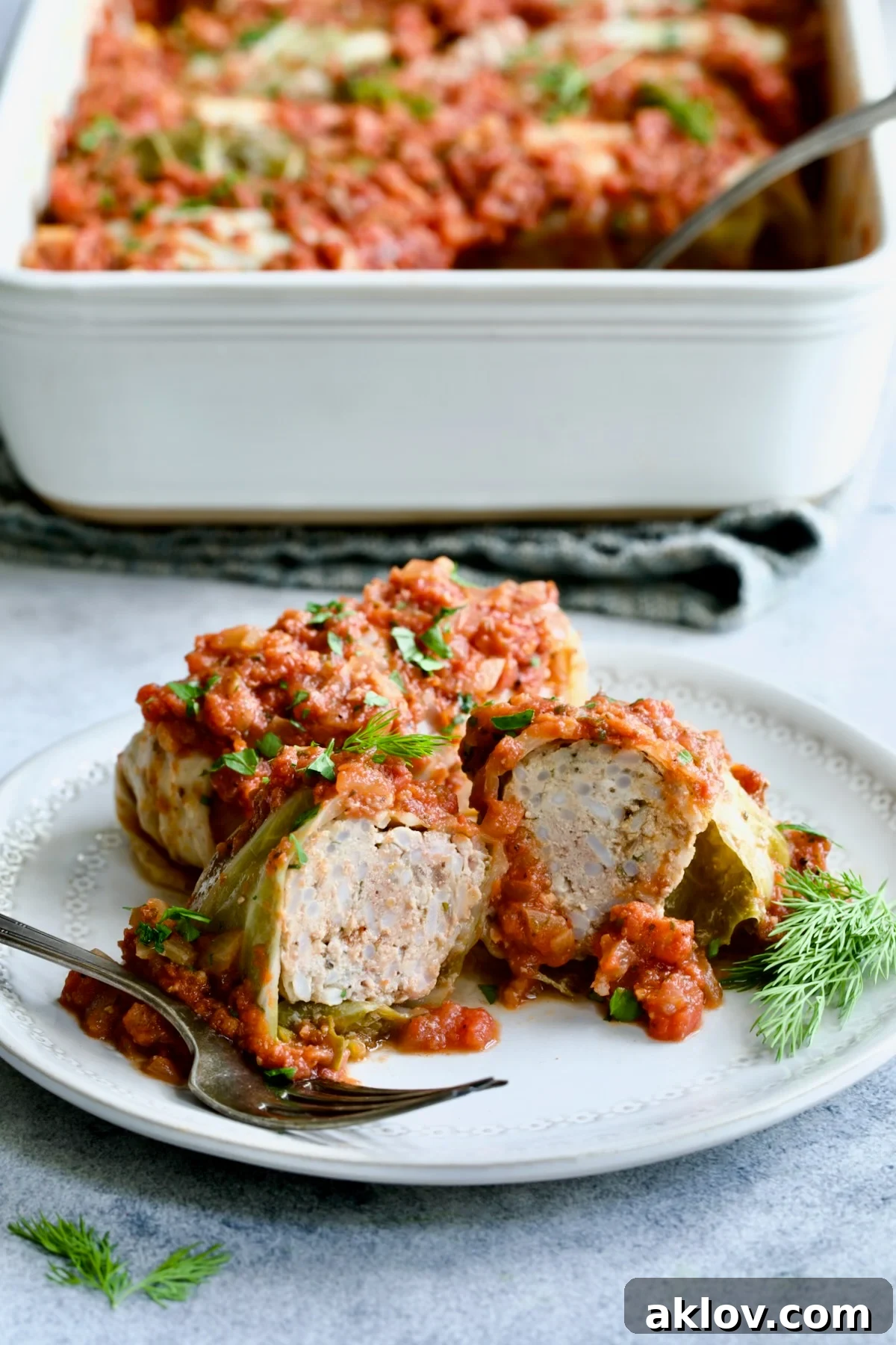 Polish Stuffed Cabbage roll cut in half to expose filling on white plate with baking dish of cabbage in the background.
