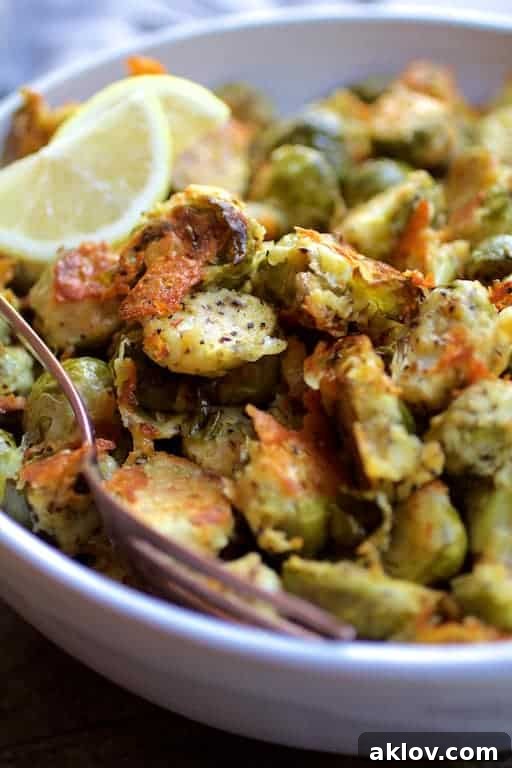 Close-up photo of Lemon Parmesan Crusted Brussels Sprouts in white bowl garnished with lemon wedges.