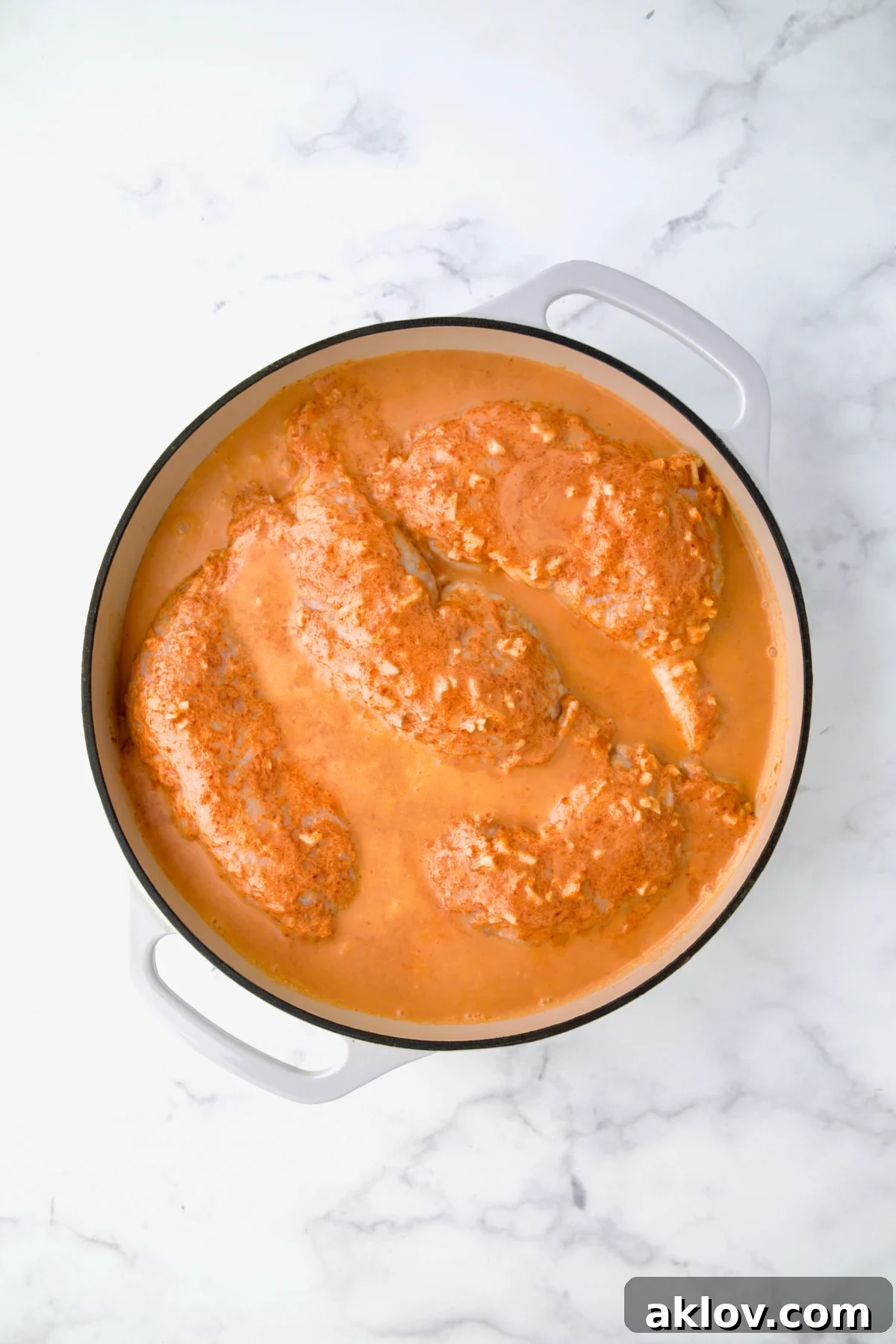 The rich coconut milk curry mixture being poured generously over seasoned chicken legs nestled in a pristine white enamel cast iron skillet, ready for braising.