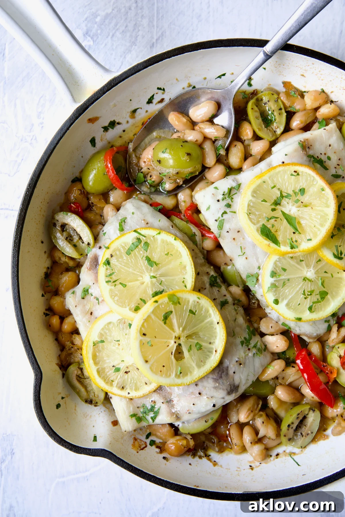 Mediterranean Fish and White Bean Skillet in white cast iron skillet topped with lemon slices with serving spoon.