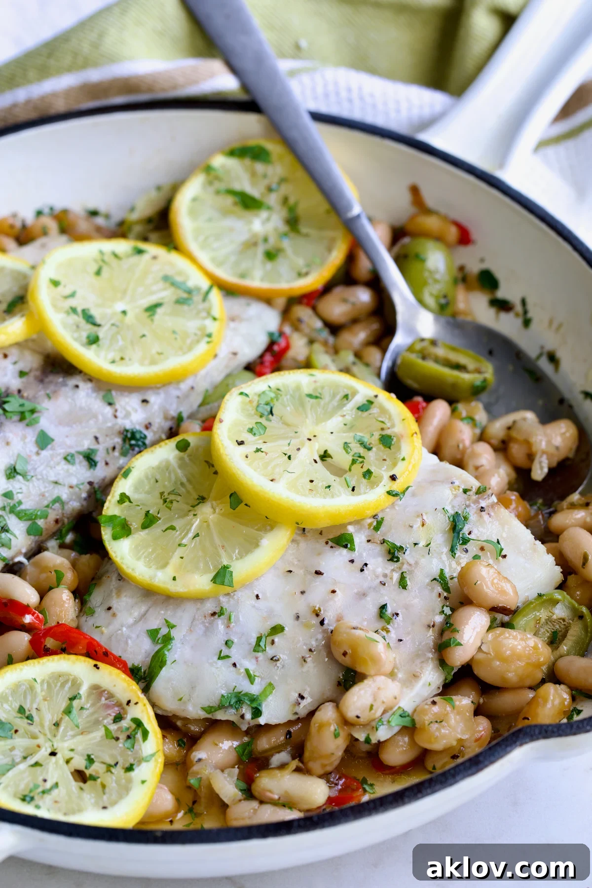 Mediterranean Fish and White Bean Skillet in white cast iron skillet topped with lemon slices with serving spoon.