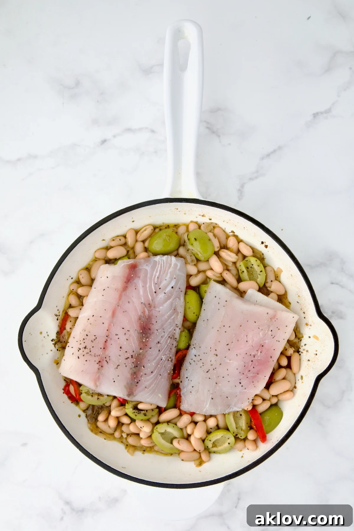 Uncooked fish fillets placed atop white bean mixture in white cast iron skillet.