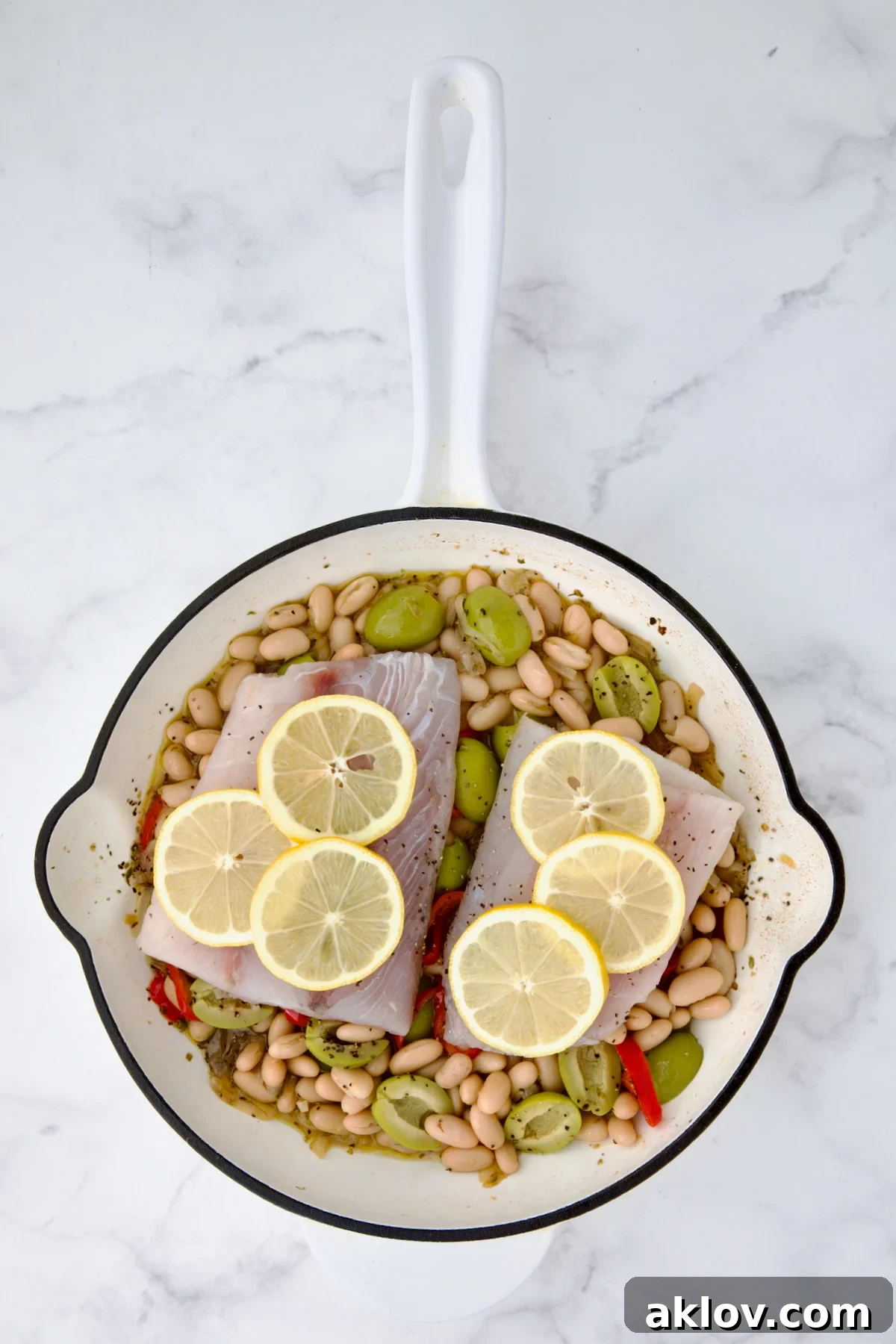 Uncooked fish fillets placed atop white bean mixture in white cast iron skillet with lemon slices added.