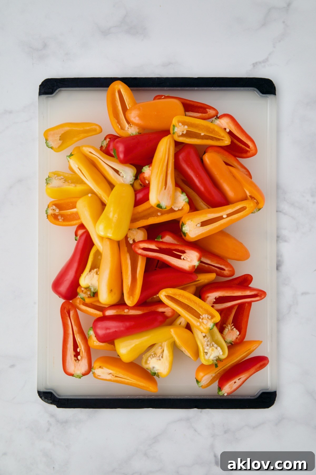 Pile of halved yellow and red mini sweet peppers on a white plastic cutting board before seeds and membranes are removed.