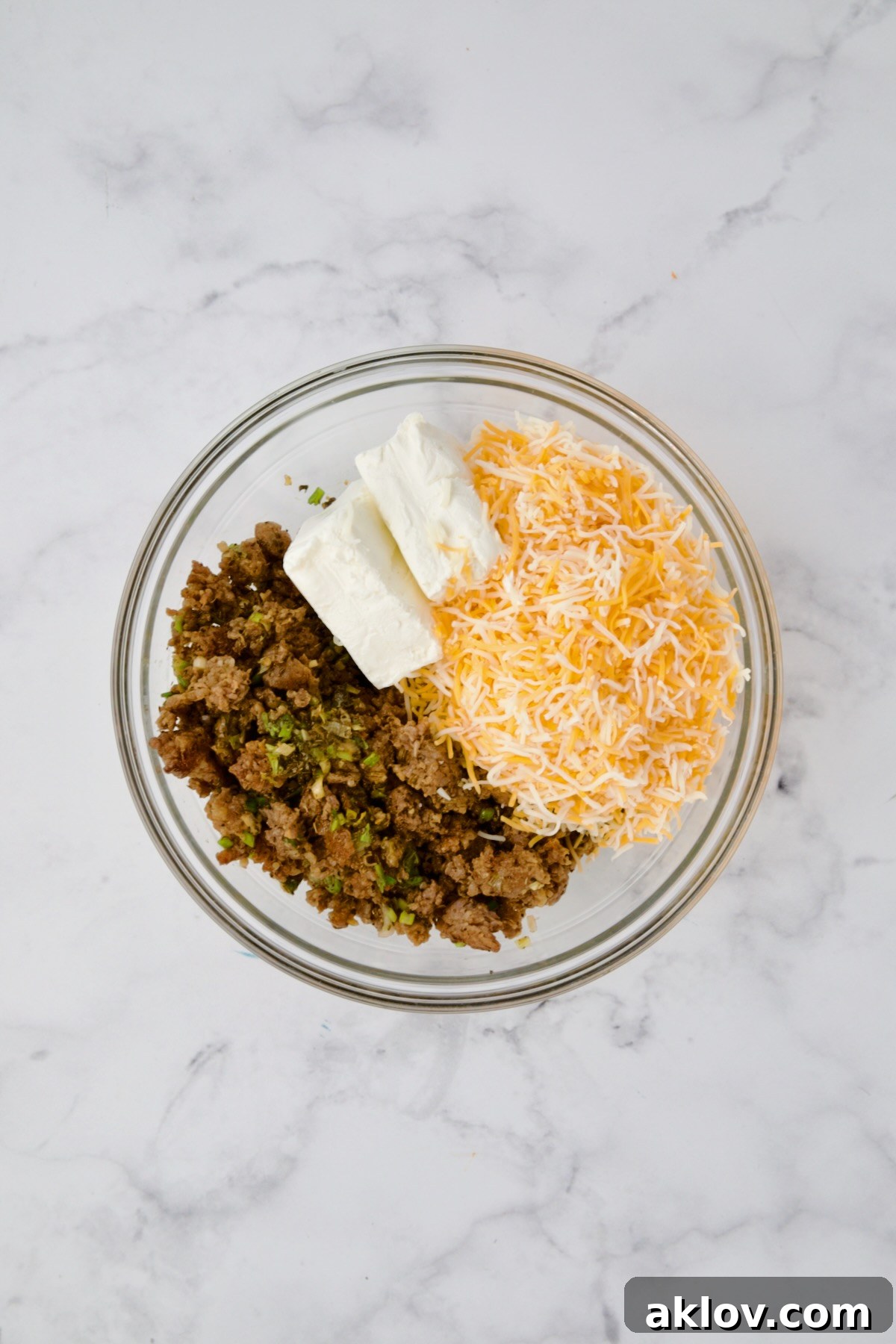 A glass bowl containing cooked sausage, softened cream cheese, and shredded Mexican blend cheese before being mixed together.