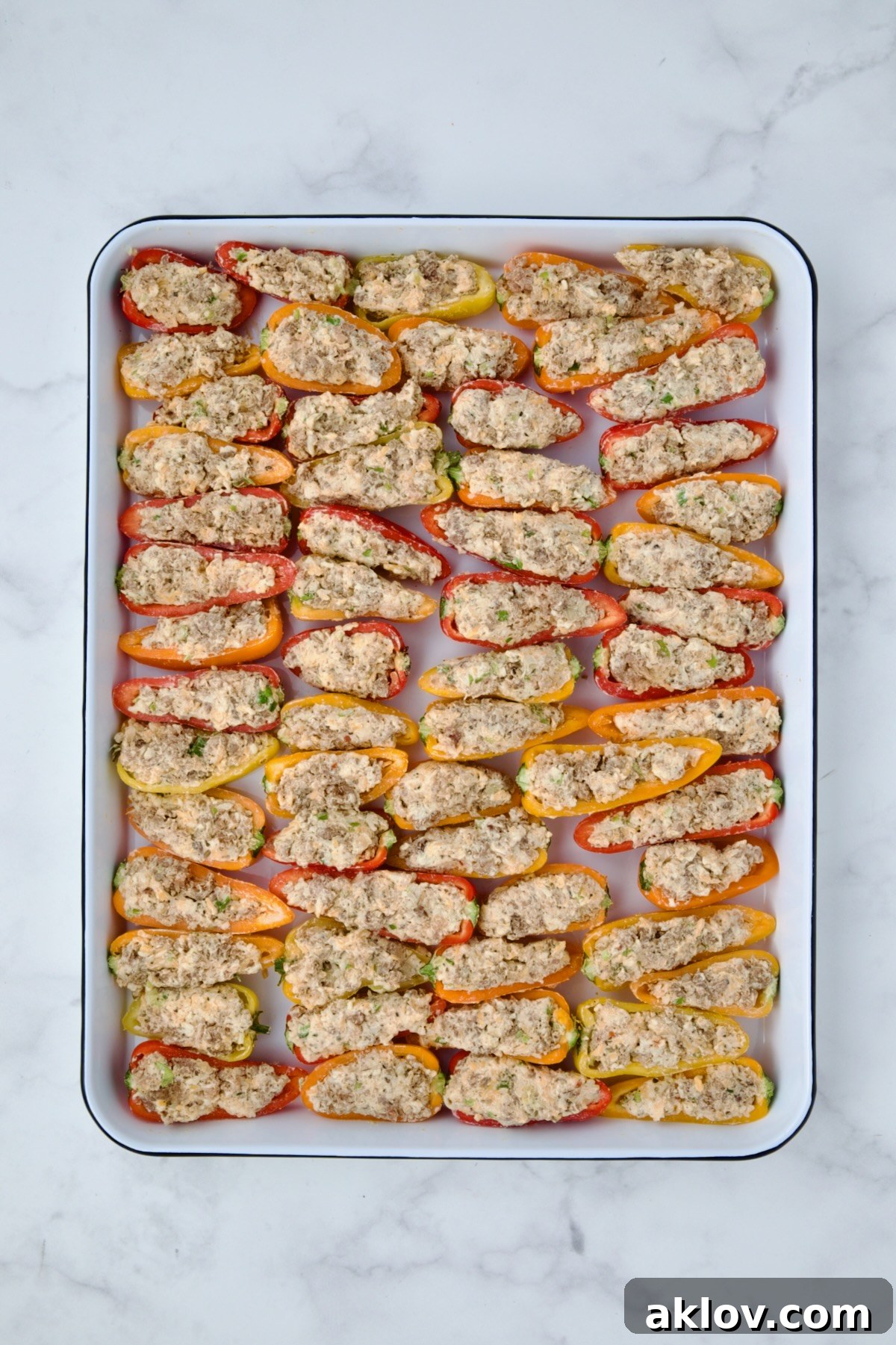 Mini peppers filled with sausage and cheese mixture arranged on a white enamel rimmed baking sheet before baking.