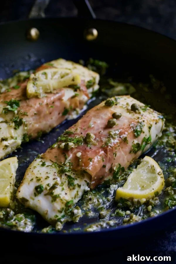 Close-up photo of Prosciutto-Wrapped Halibut with Herb-Caper Butter and lemon wedges in a black skillet, ready to be served.