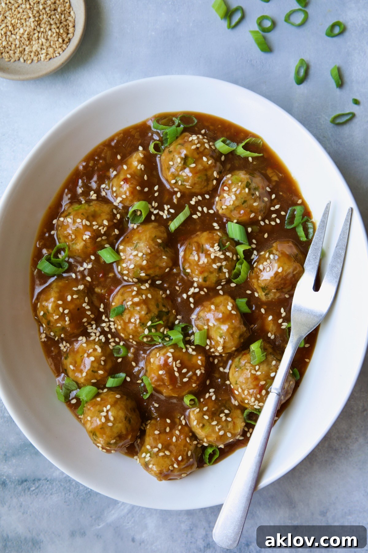 Plated Asian Turkey Meatballs in a white oval serving bowl, beautifully garnished with toasted sesame seeds and sliced green scallions.