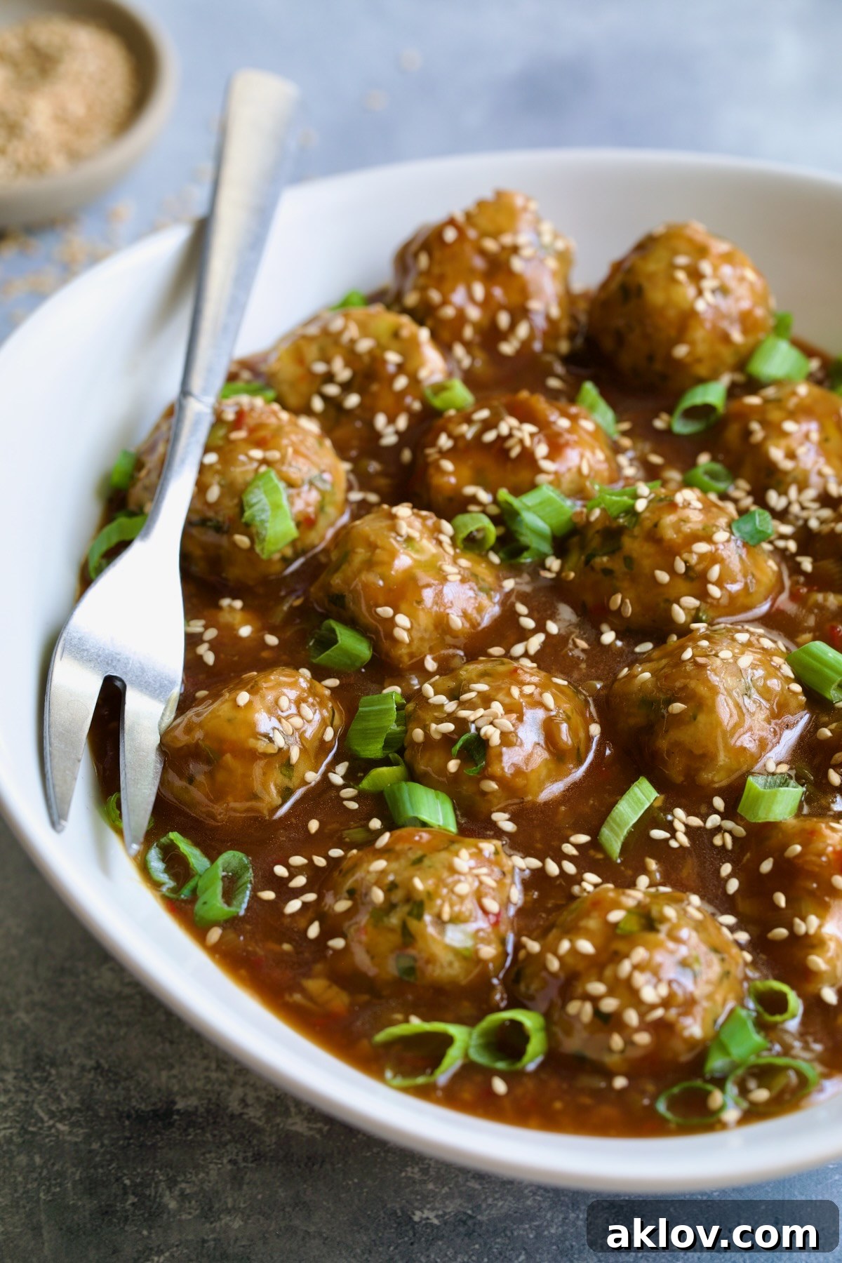 Close-up view of Asian Turkey Meatballs in a white oval serving dish, adorned with sesame seeds and green scallions, with a serving fork.