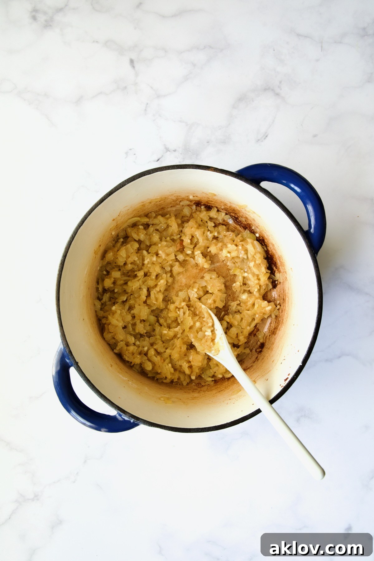 Flour fully incorporated into the cooked onions and garlic, forming a fragrant roux in a blue Dutch oven, stirred with a white mixing spoon.