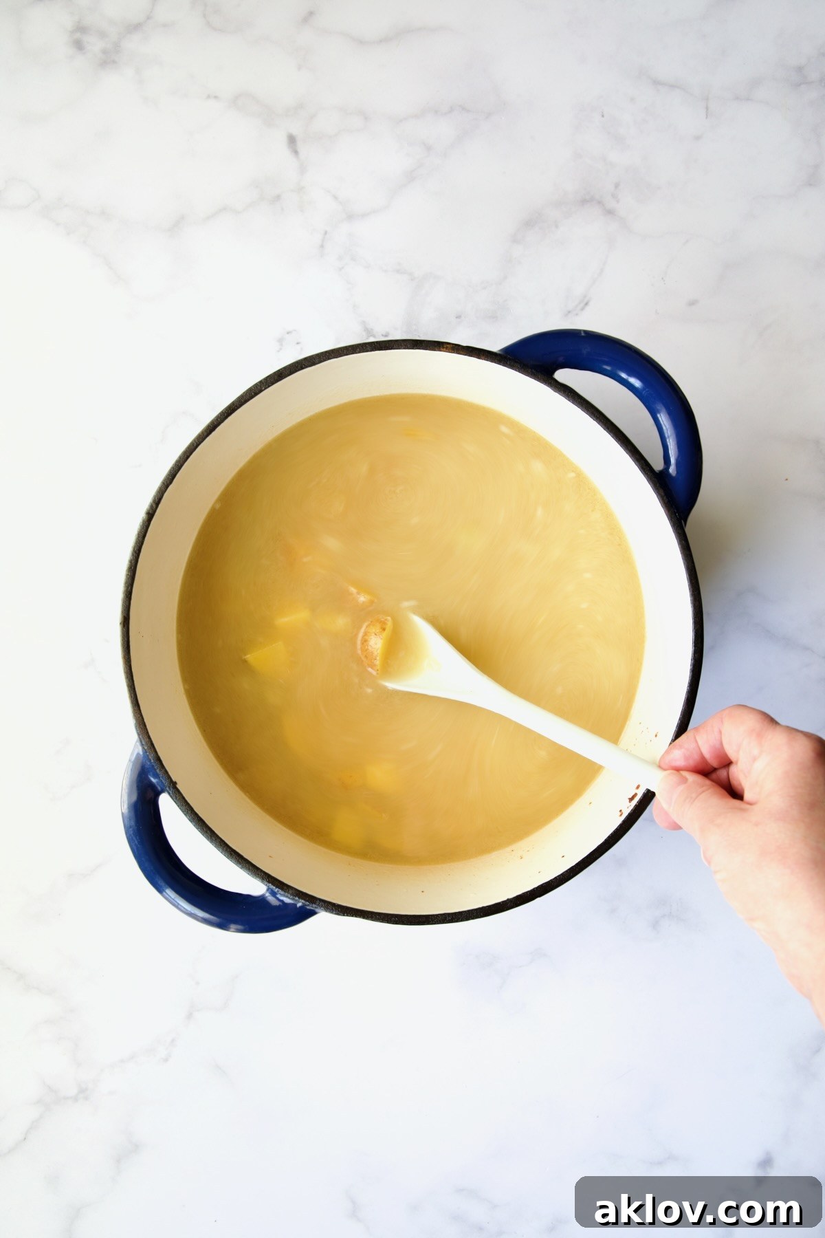 Chicken broth and diced potatoes being added to the roux in a blue Dutch oven, with a hand stirring the mixture with a white spoon.