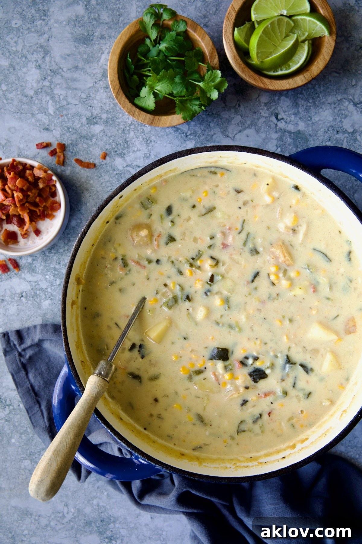 The finished Roasted Poblano Corn Chowder presented in a beautiful blue Dutch oven, accompanied by a ladle and small bowls containing garnishes like bacon, cilantro, and lime wedges.