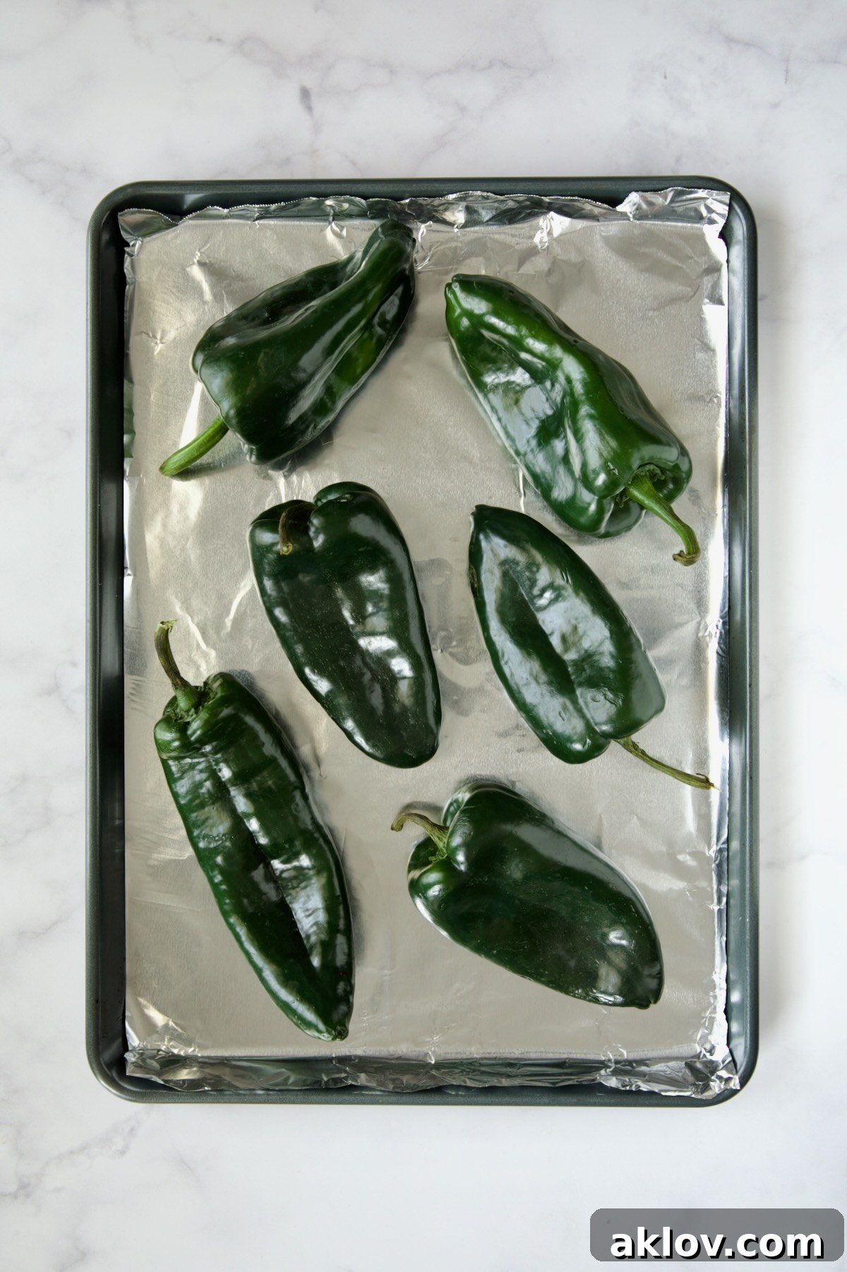 Six fresh, unroasted poblano peppers neatly arranged on an aluminum-foil lined rimmed baking sheet, ready for roasting.