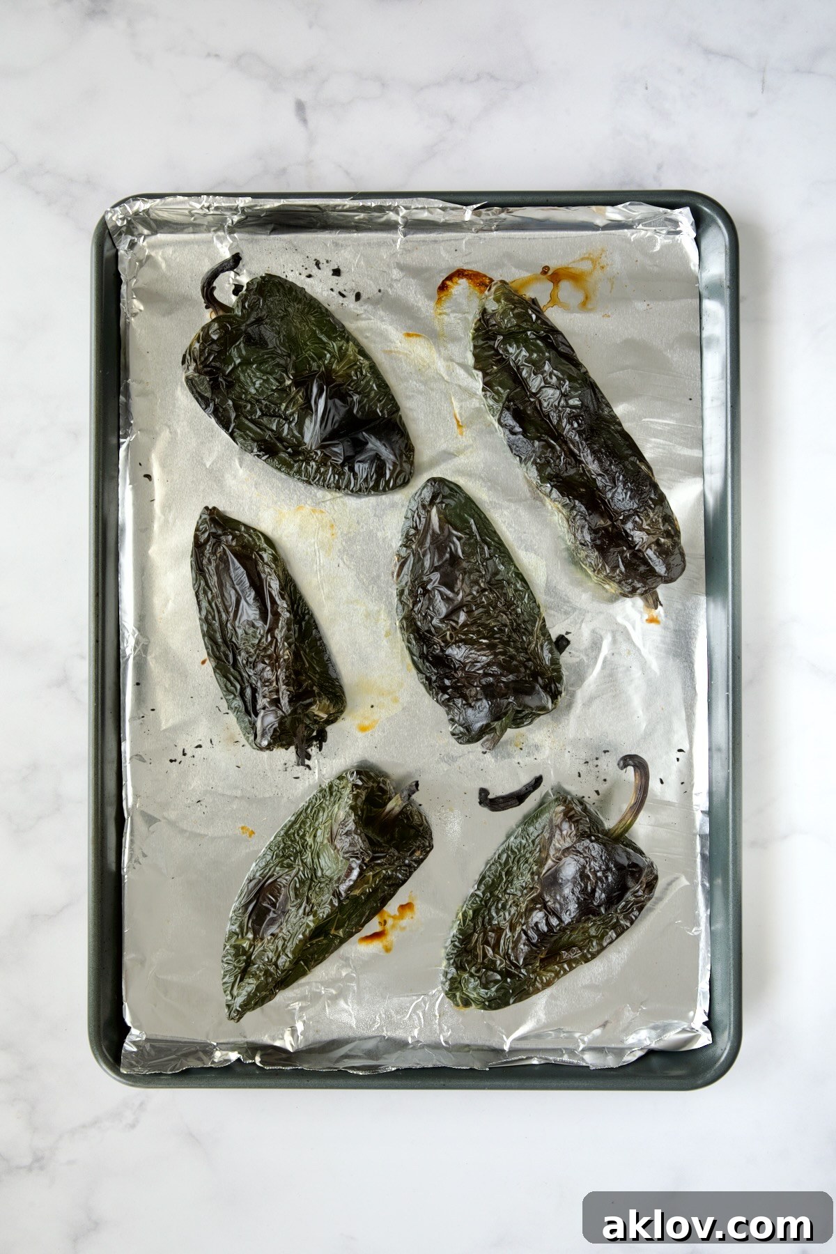 Six perfectly roasted poblano peppers, their skins blackened and blistered, resting on an aluminum foil lined rimmed baking sheet after broiling.