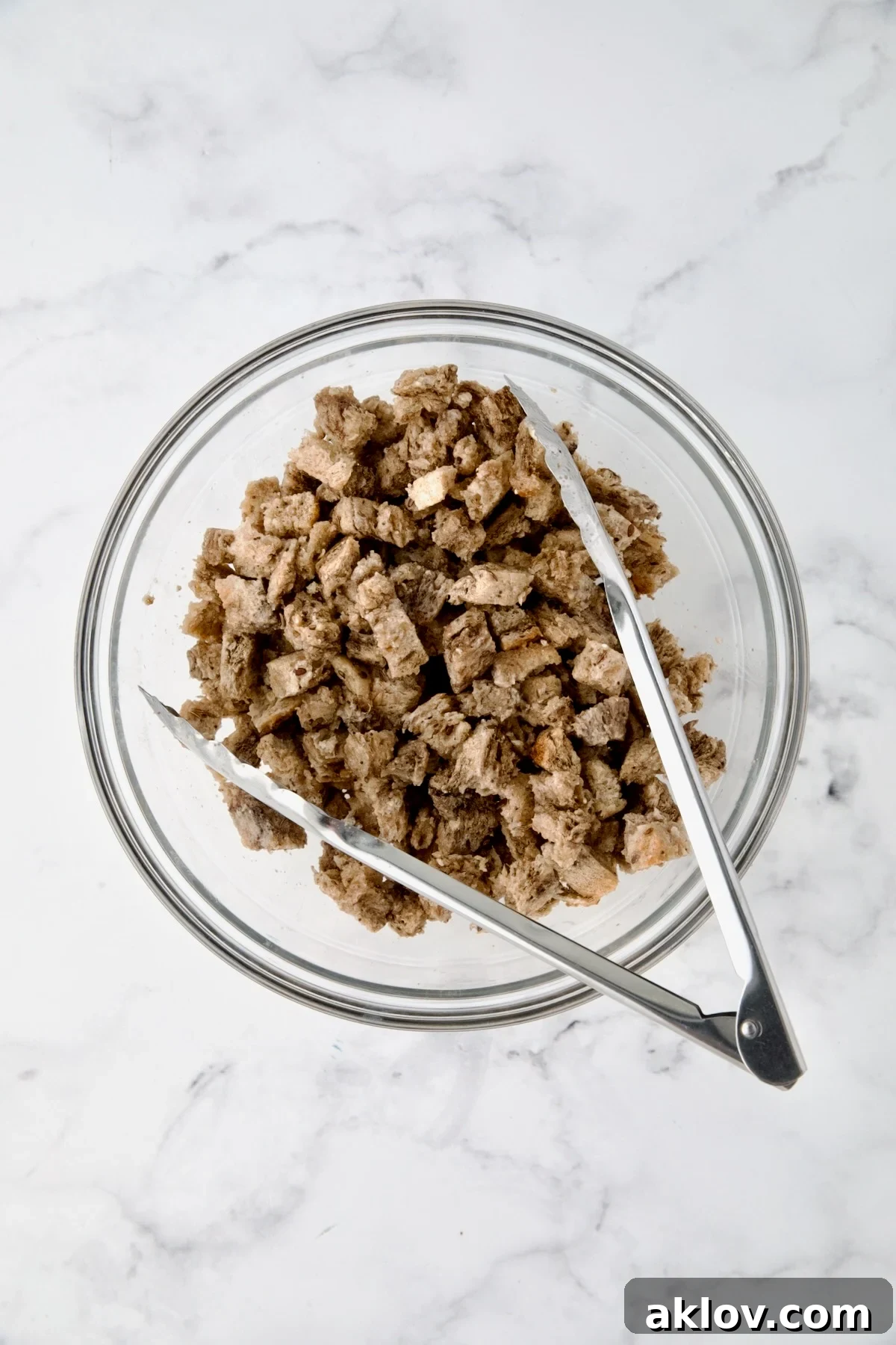 Creamy Reuben Comfort Casserole 6 Kitchen tongs gently tossing rye bread cubes with melted butter in a clear glass bowl, ensuring even coating.