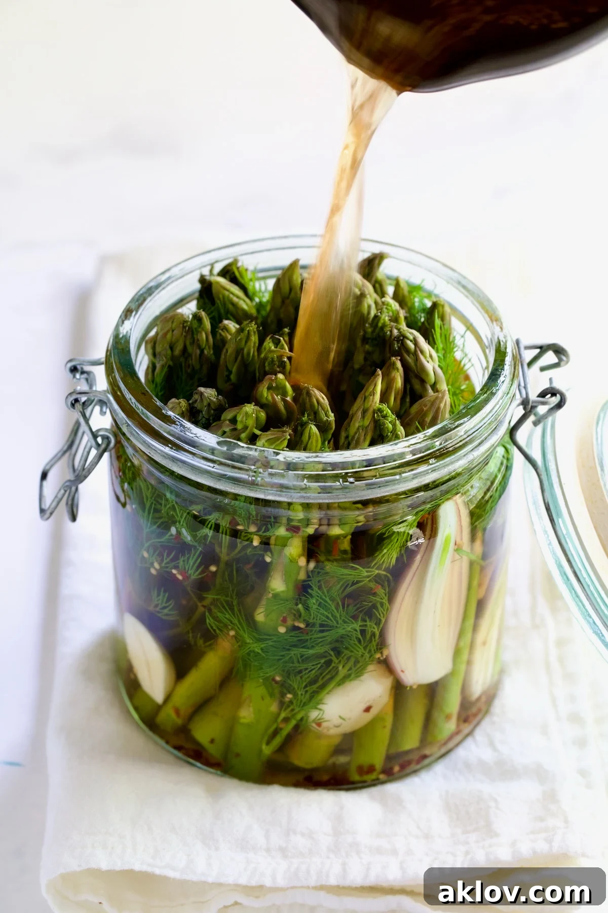 Zesty Pickled Asparagus 8 Hot pickling brine being carefully poured over the packed asparagus, garlic, shallot, and dill in a glass jar, ensuring all vegetables are fully submerged.