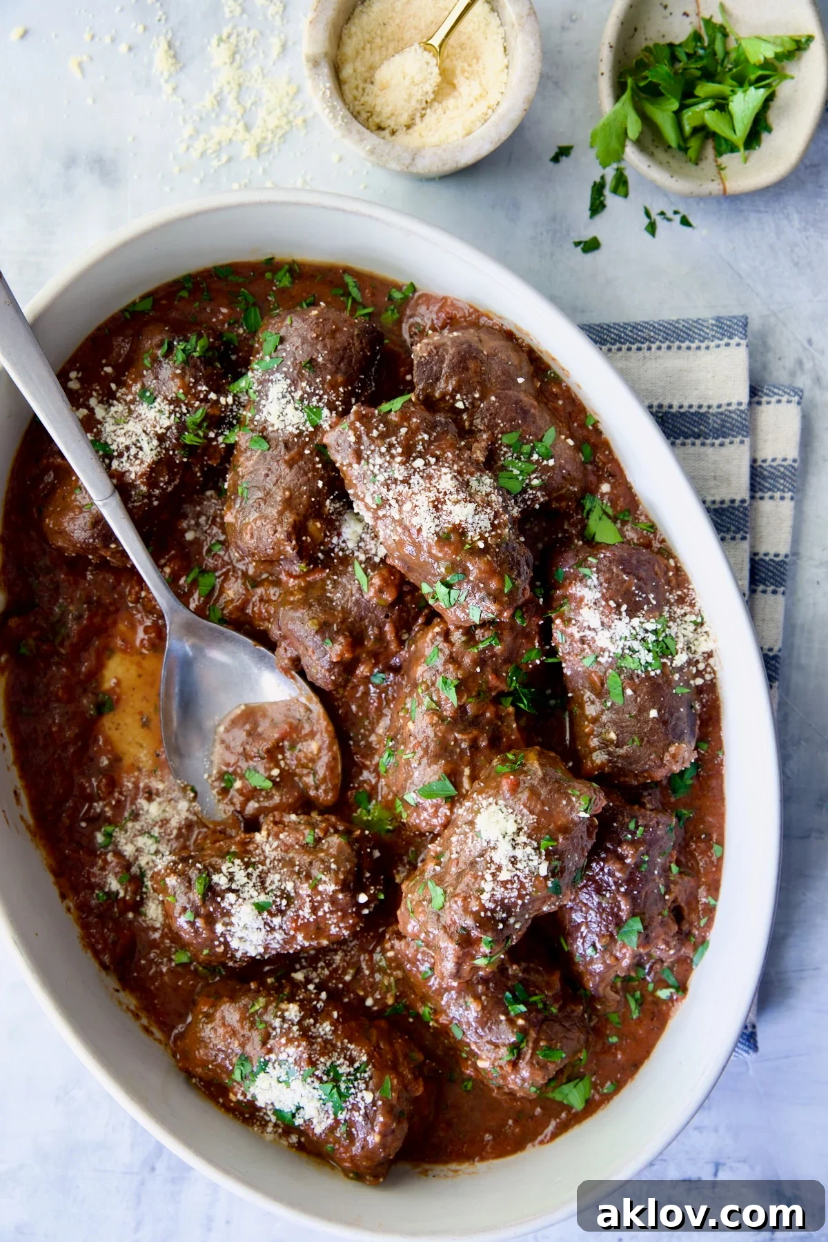 Beef Braciole in oval white serving dish with serving spoon on blue striped towel.