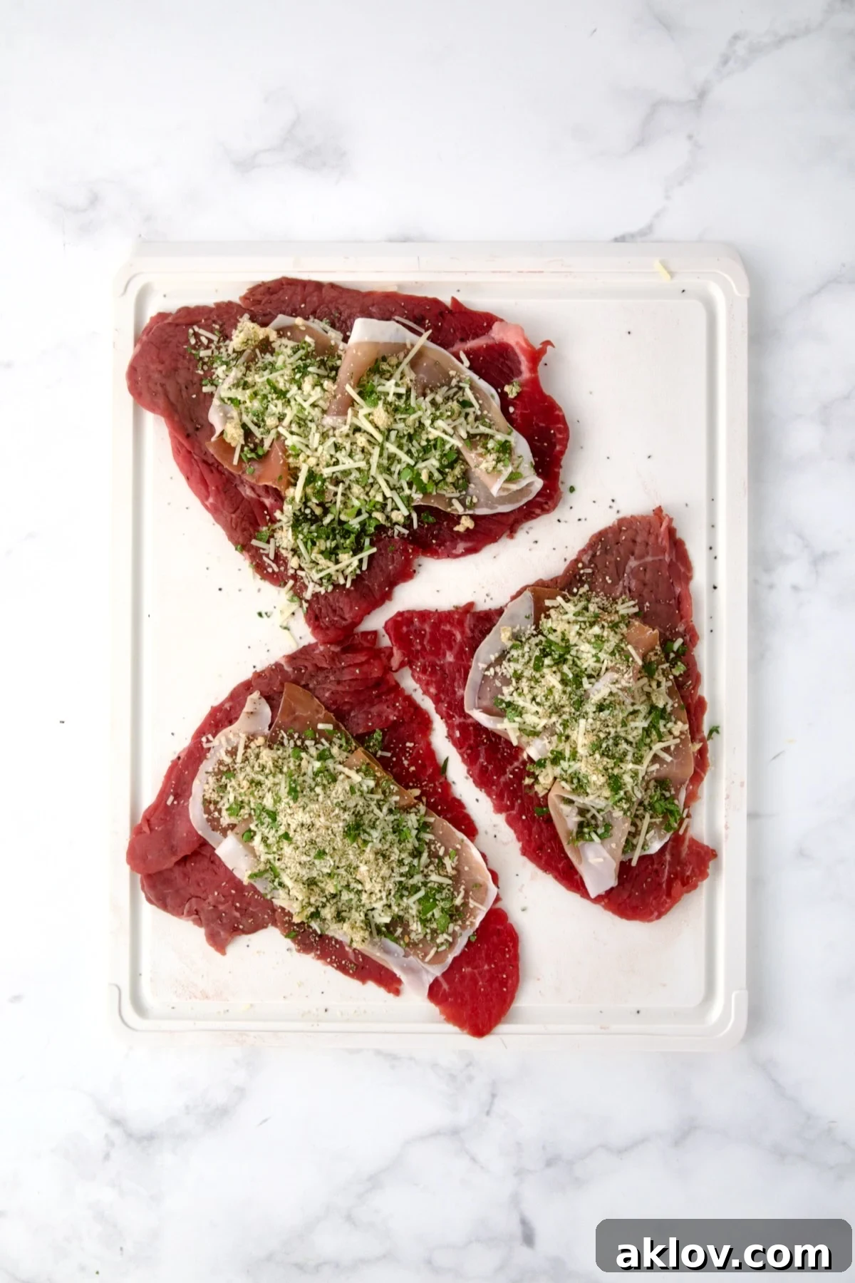 Three slices of beef top round on white cutting board with slices of prosciutto and the filling placed on top of the slices.