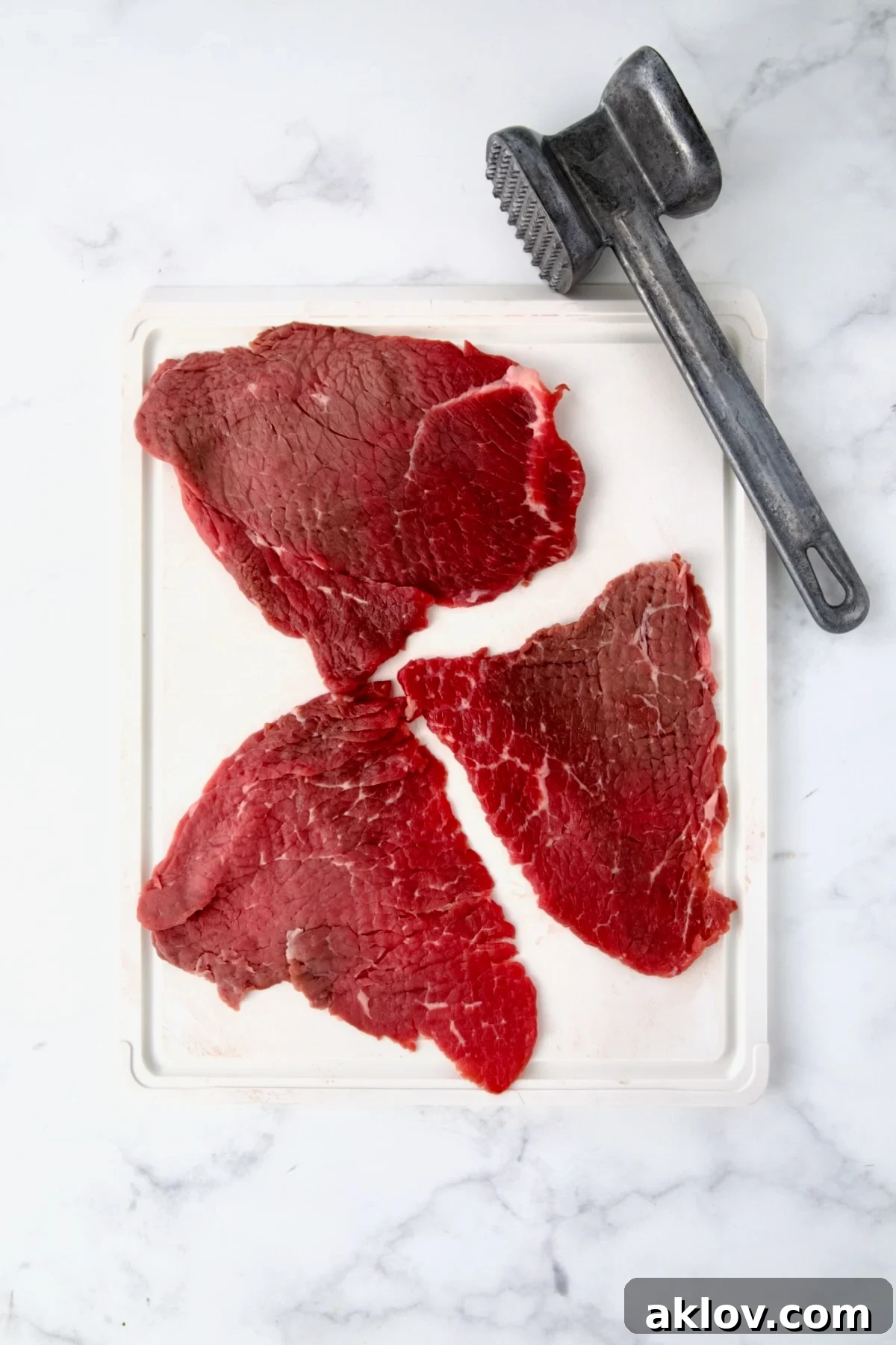 Three slices of beef top round on white cutting board with meat mallet to show how it's pounded.