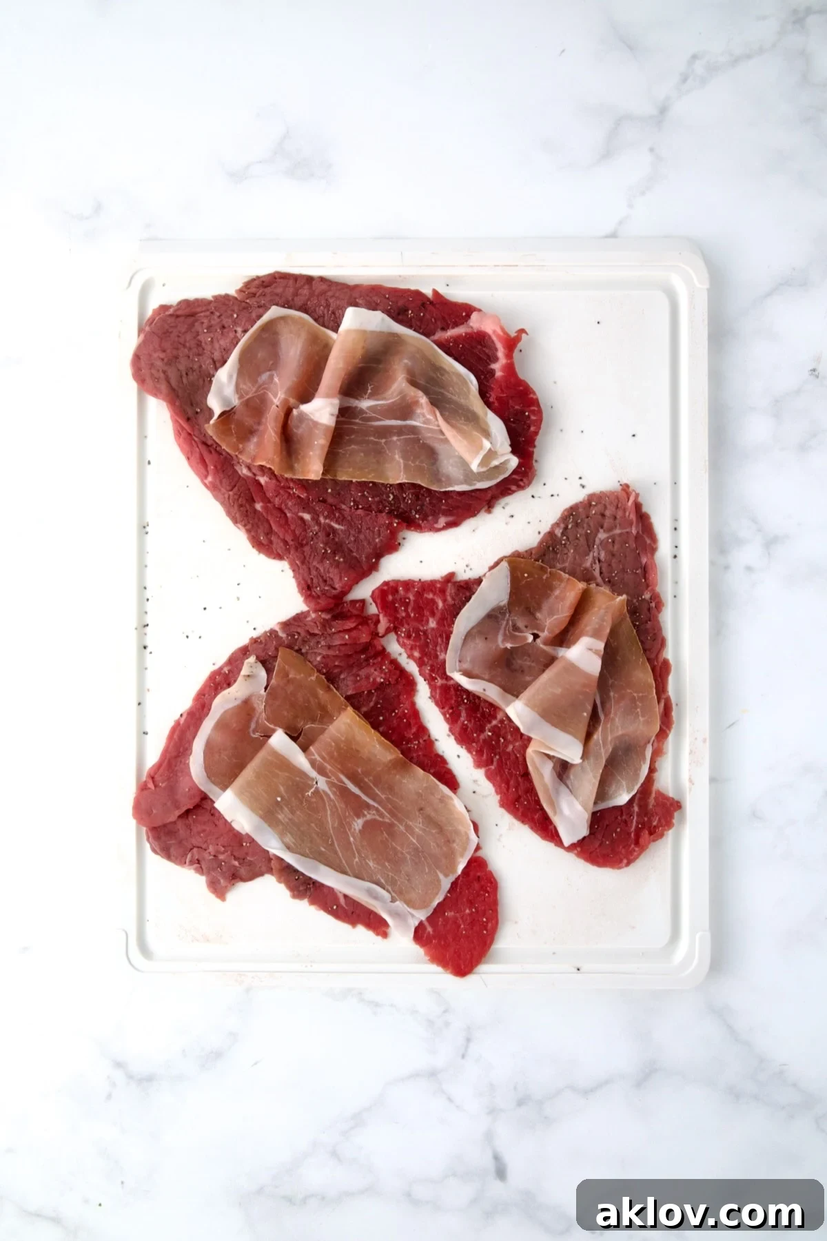 Three slices of beef top round on white cutting board seasoned with salt and black pepper with slices of prosciutto placed on top of the slices.
