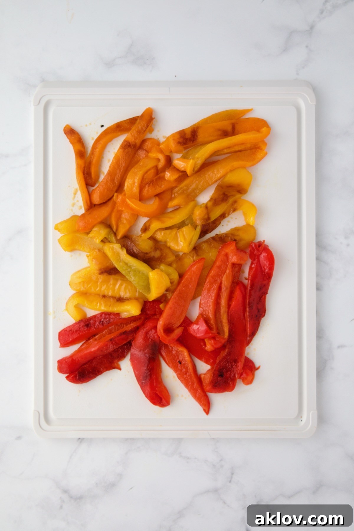 Orange, yellow and red bell pepper halves cut into strips on white plastic cutting board.