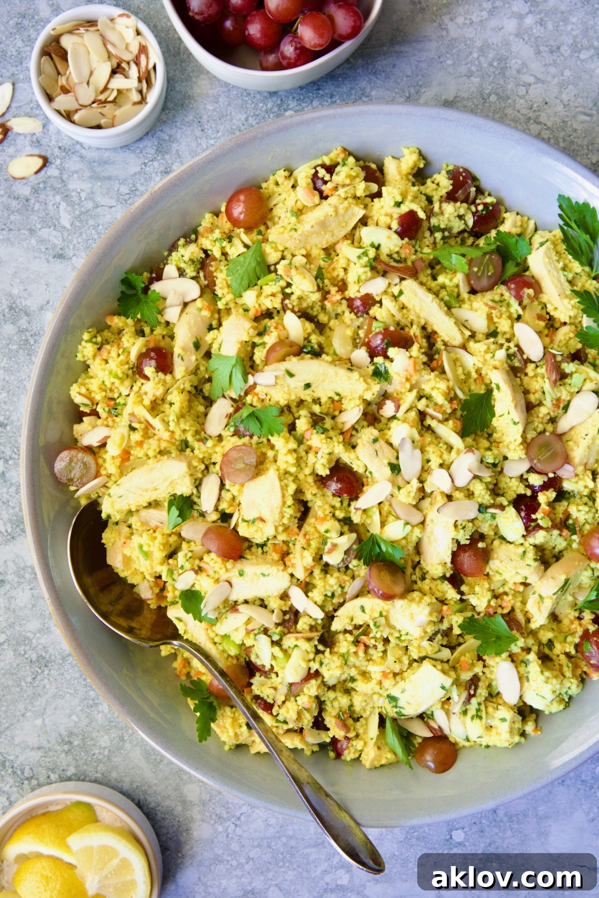Chicken Couscous Salad in a sleek gray bowl with a serving spoon, surrounded by small bowls of sliced almonds, whole grapes, and fresh lemon wedges, highlighting the salad's vibrant ingredients.