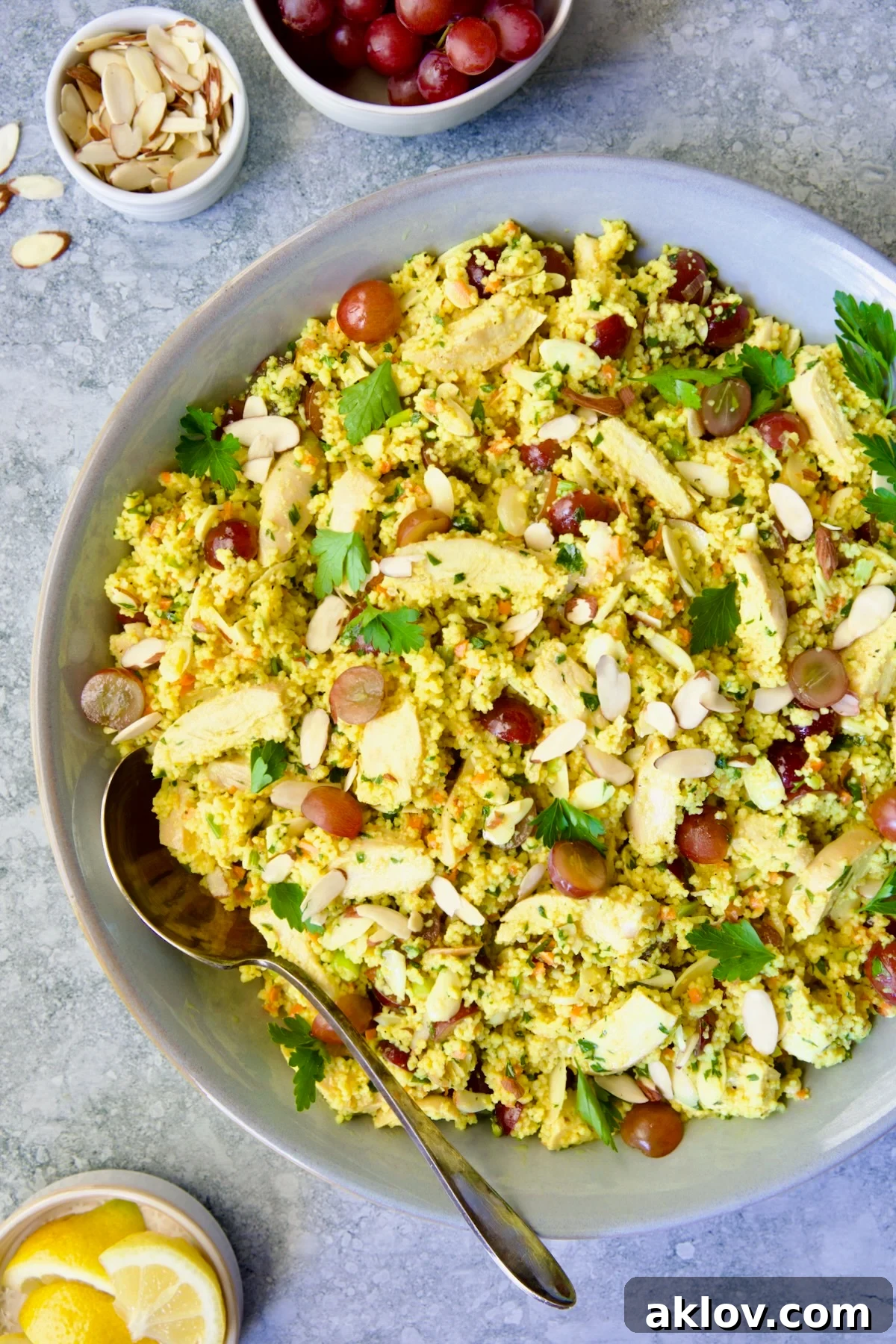 Chicken Couscous Salad in gray bowl with serving spoon with bowl of sliced almonds, whole grapes and lemon wedges.