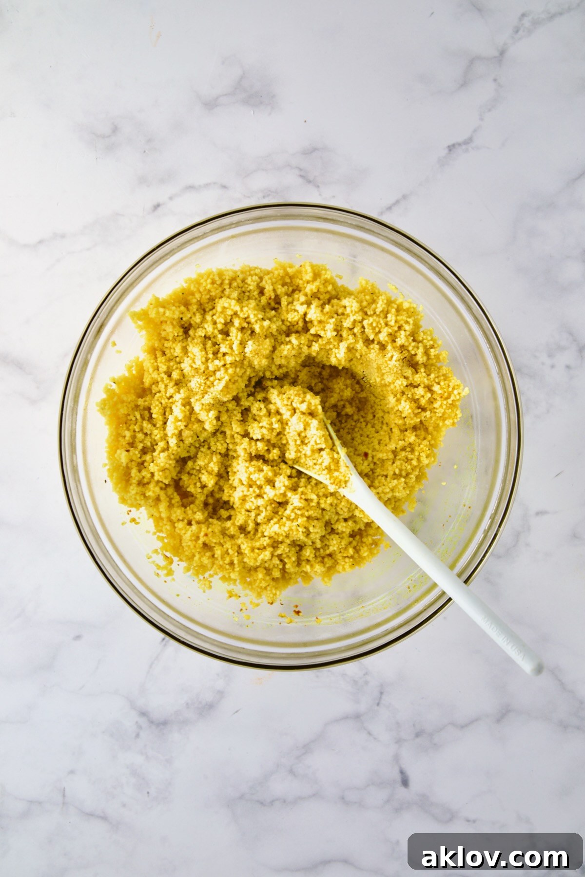 Perfectly cooked and fluffed couscous in a glass bowl, being gently separated with a white mixing spoon, indicating readiness for the salad.