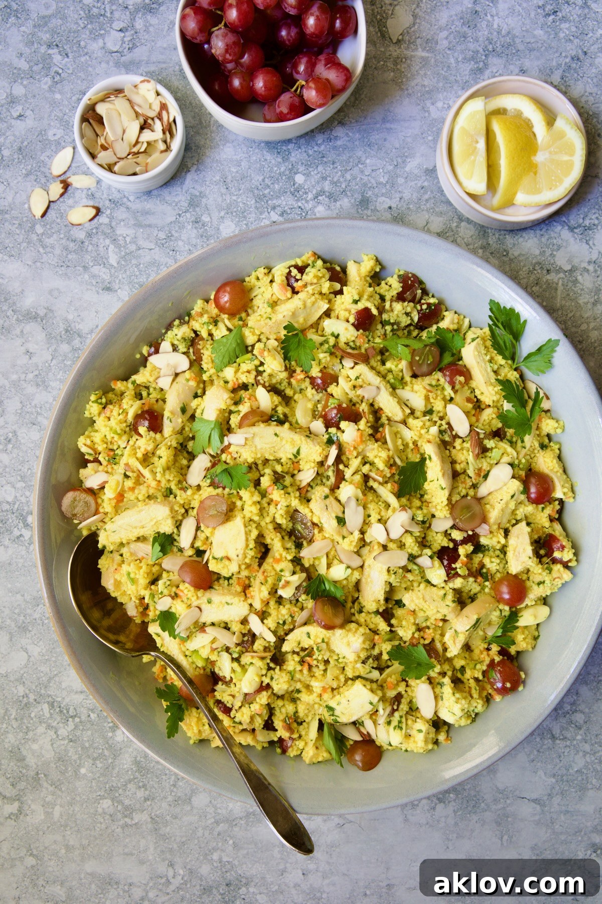 A beautifully presented Chicken Couscous Salad in a gray serving bowl with a spoon, accompanied by small dishes of extra sliced almonds, whole grapes, and lemon wedges for garnish.
