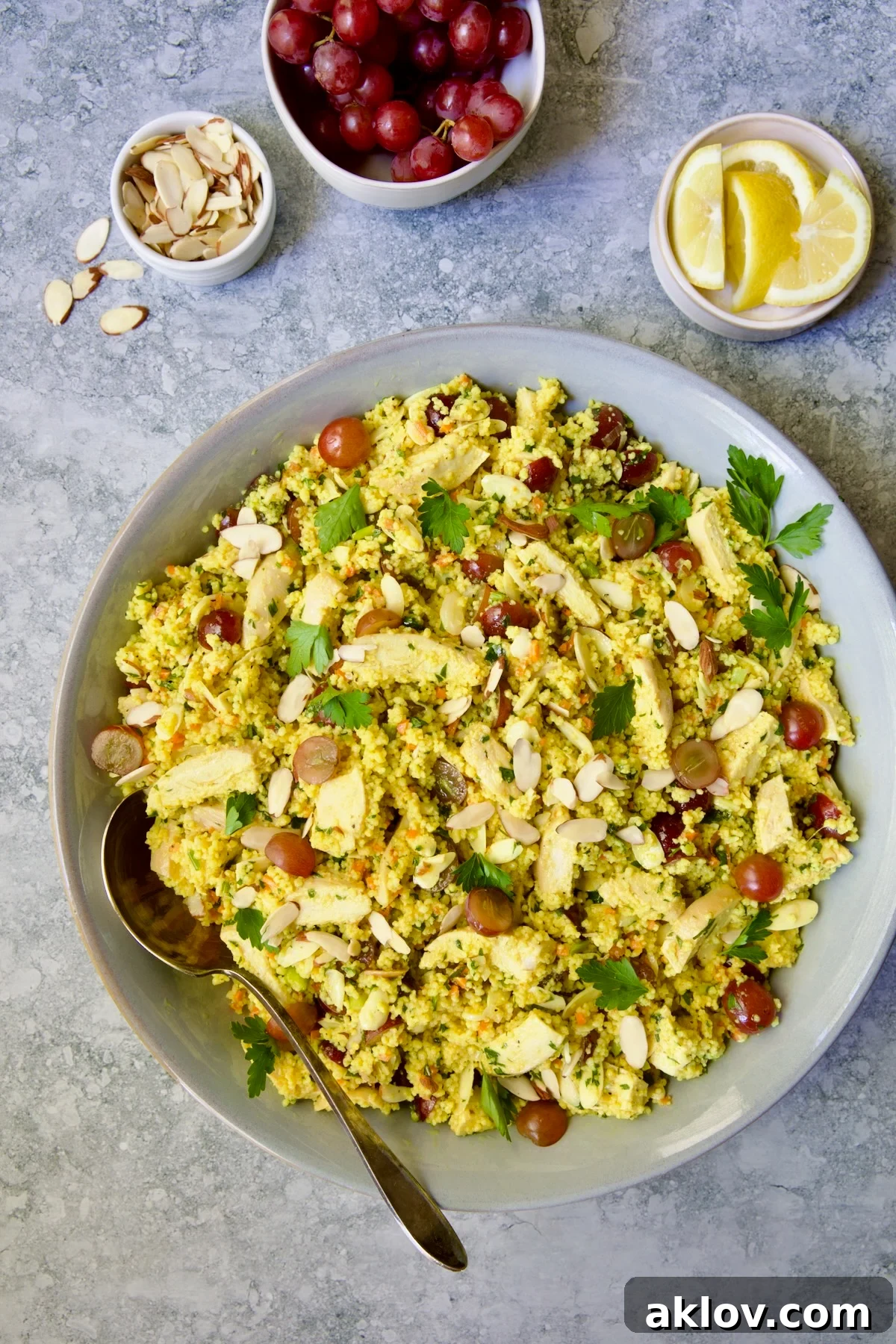 Chicken Couscous Salad in gray bowl with serving spoon with bowl of sliced almonds, whole grapes and lemon wedges.