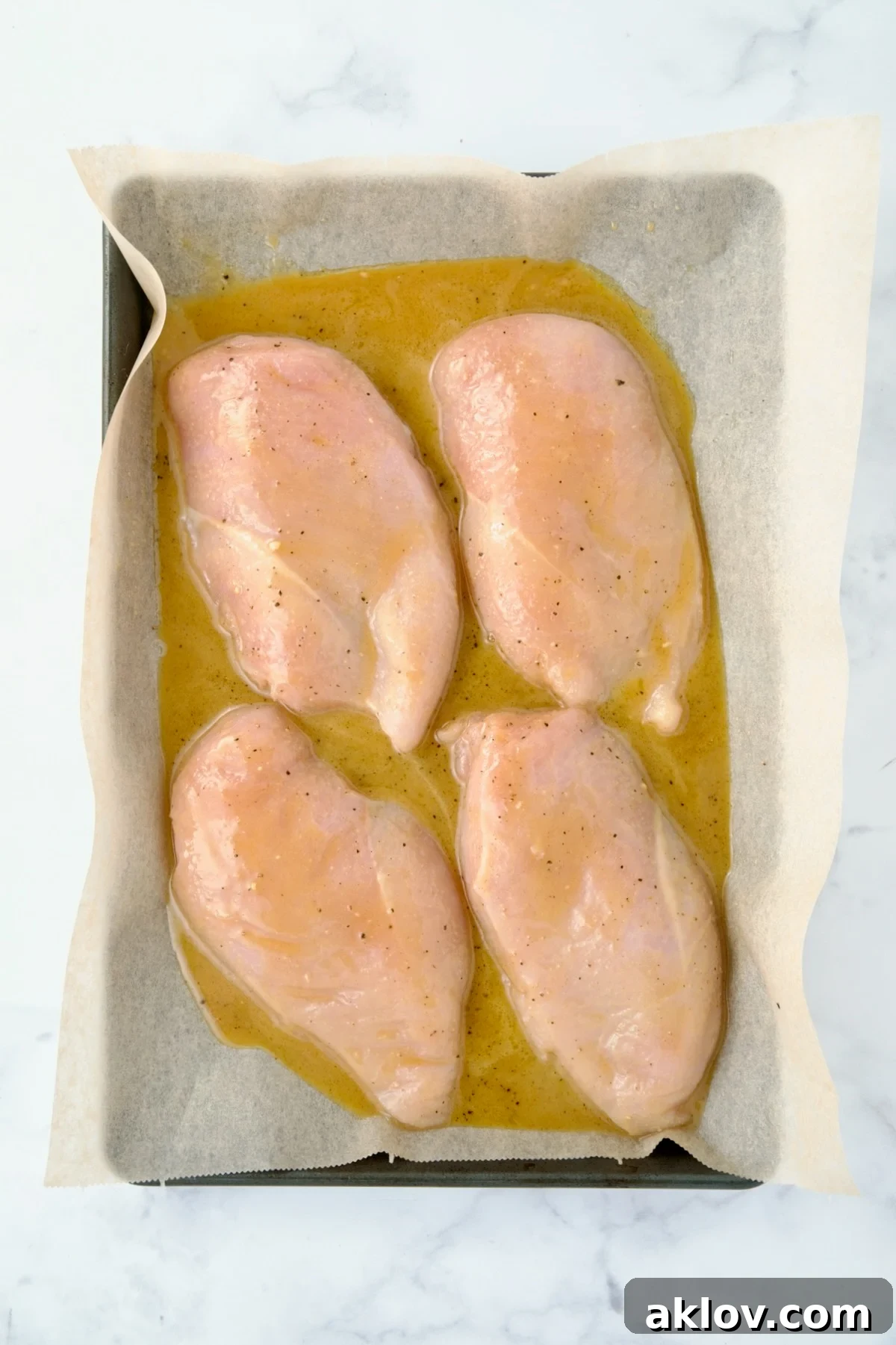 Four uncooked chicken breast halves on parchment paper lined rimmed baking sheet with honey mustard combination poured over them.